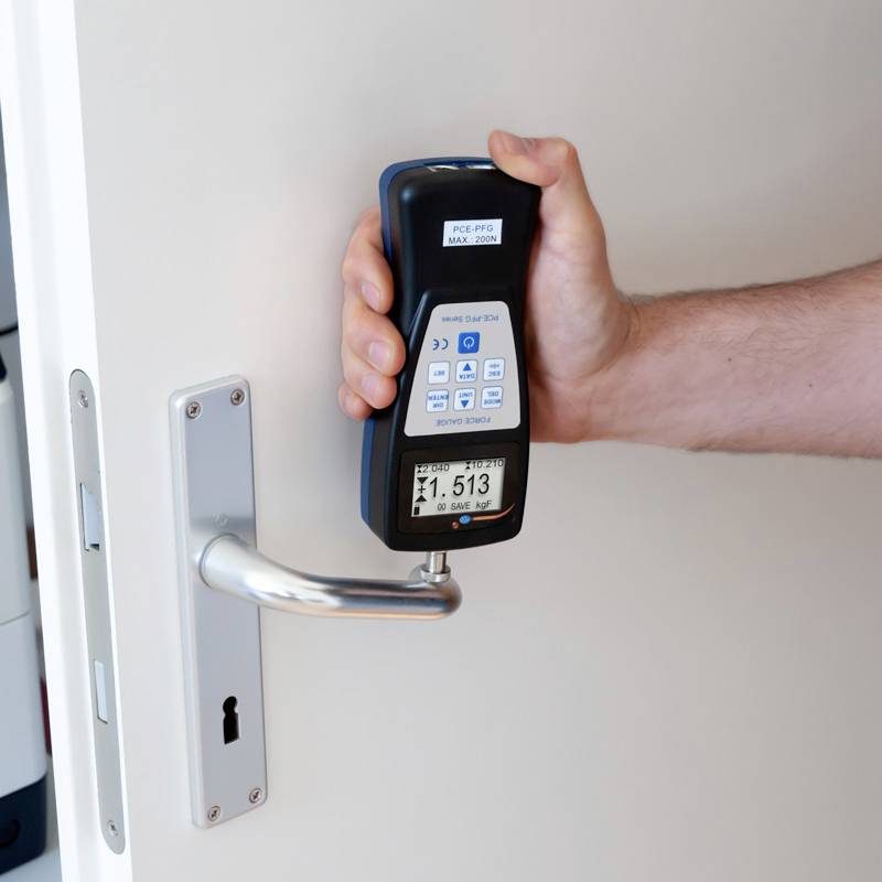 Une personne tient un appareil de mesure contre une porte pour tester les vibrations. L'appareil affiche des valeurs de mesure sur un écran.