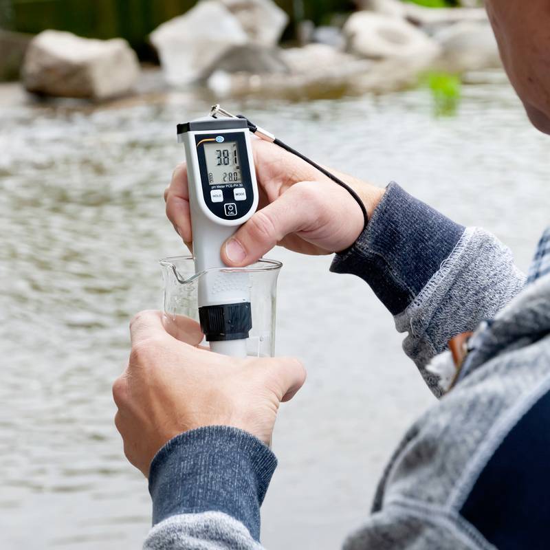 Une personne mesure la température de l'eau d'un étang à l'aide d'un thermomètre numérique qui indique 20,8 degrés Celsius.