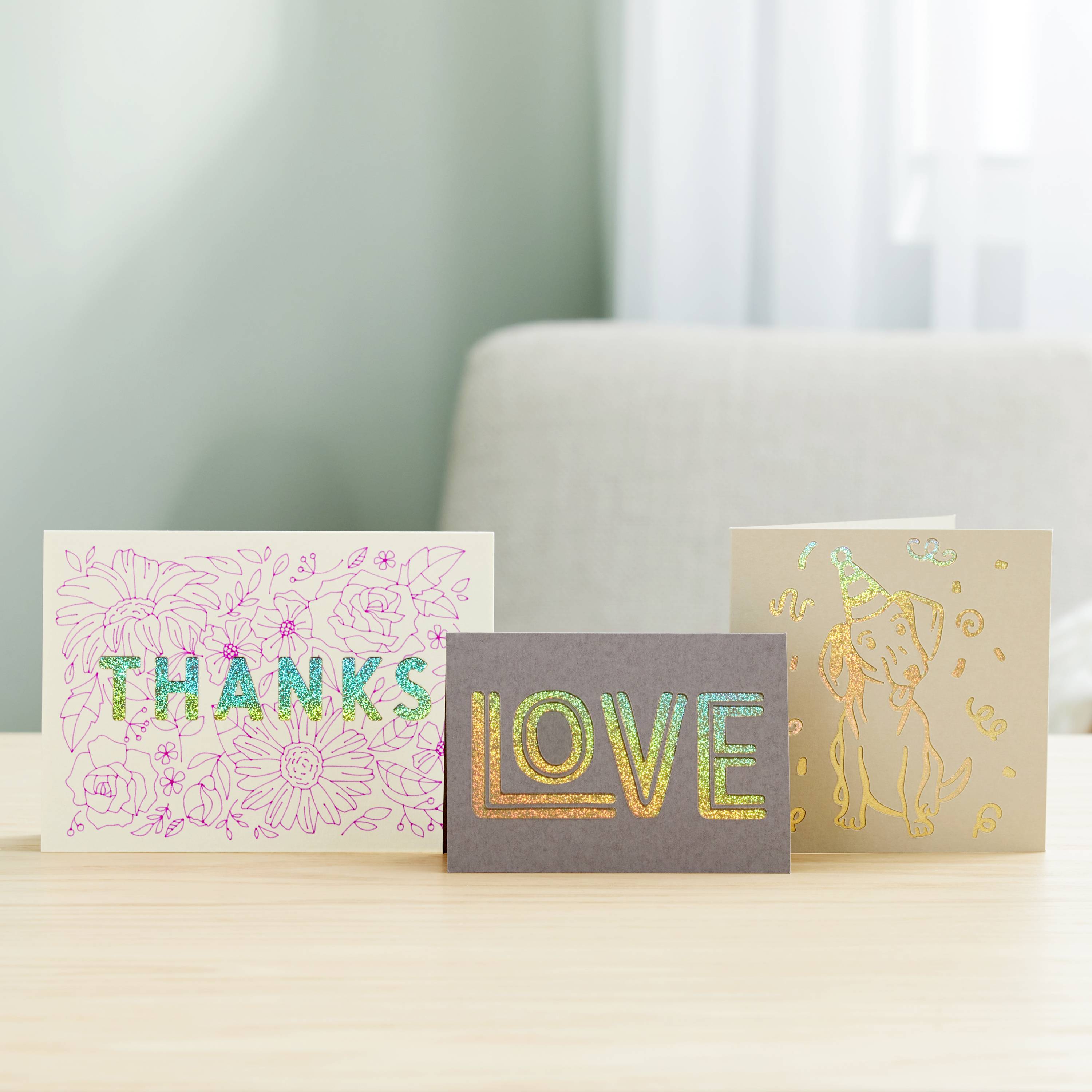 Trois cartes de vœux sur une table : à gauche 'MERCI' avec un motif floral, au centre 'AMOUR' en lettres colorées, à droite un motif de chien.