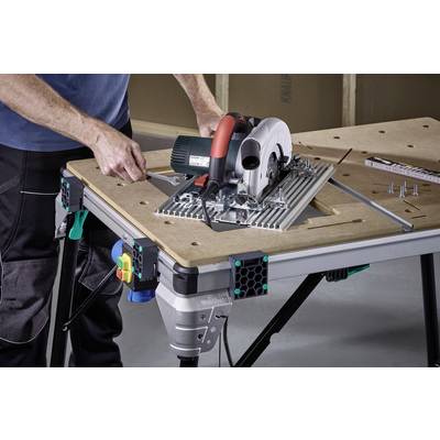 Un homme utilise une scie circulaire de table sur un établi dans un atelier. Des murs en bois sont visibles en arrière-plan, des vis sont prêtes à être utilisées.
