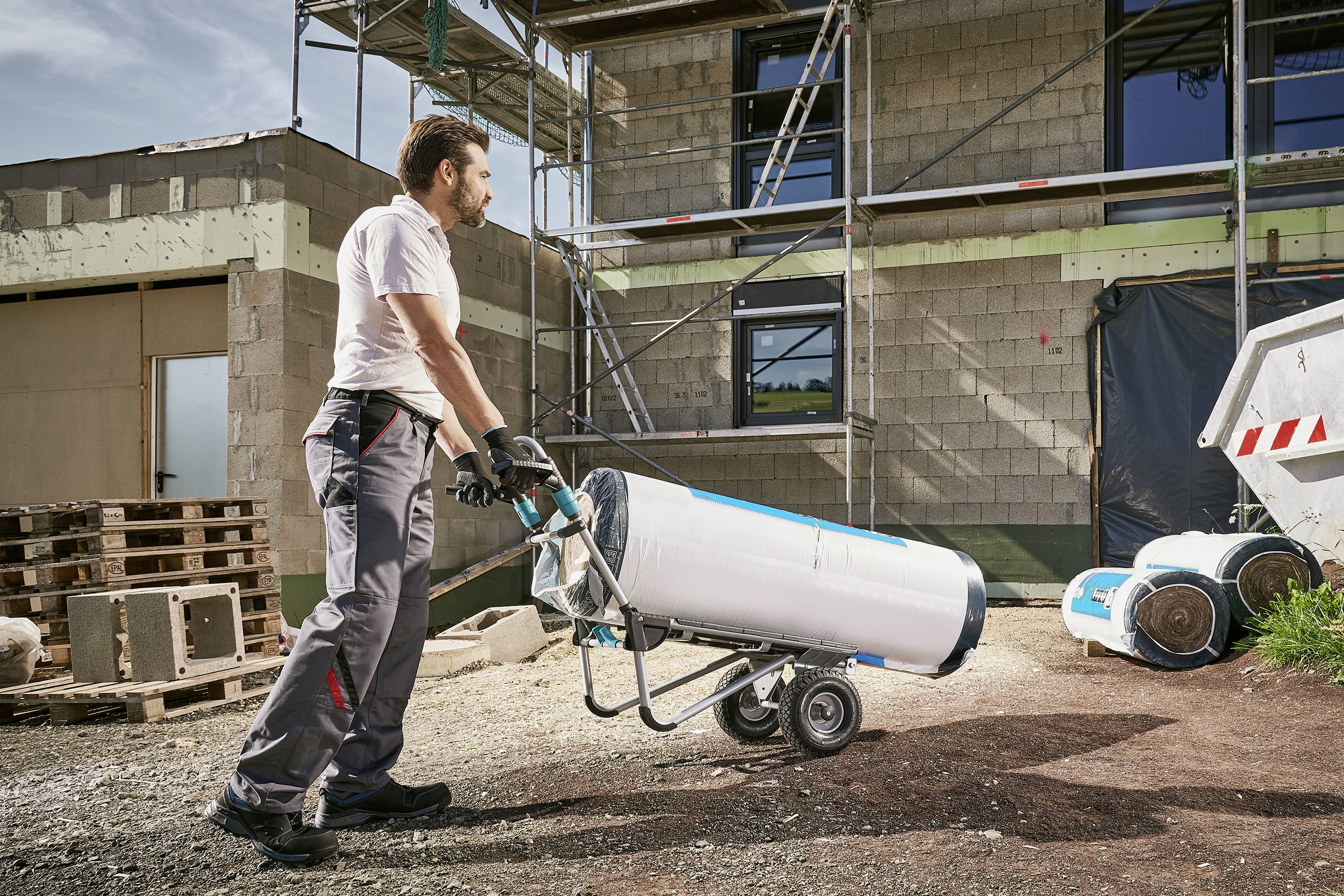 Un homme pousse un diable à roues avec un grand tonneau blanc sur un chantier de construction. Un bâtiment avec un échafaudage est visible en arrière-plan.