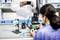 Une femme soude une carte électronique dans un atelier. Un homme en blouse blanche la photographie avec un smartphone.