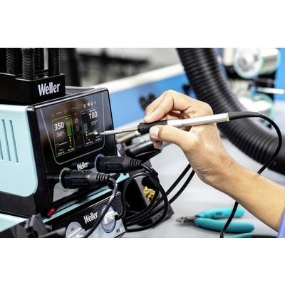 Une personne tient un fer à souder connecté à une station de contrôle de Weller sur une table de travail.