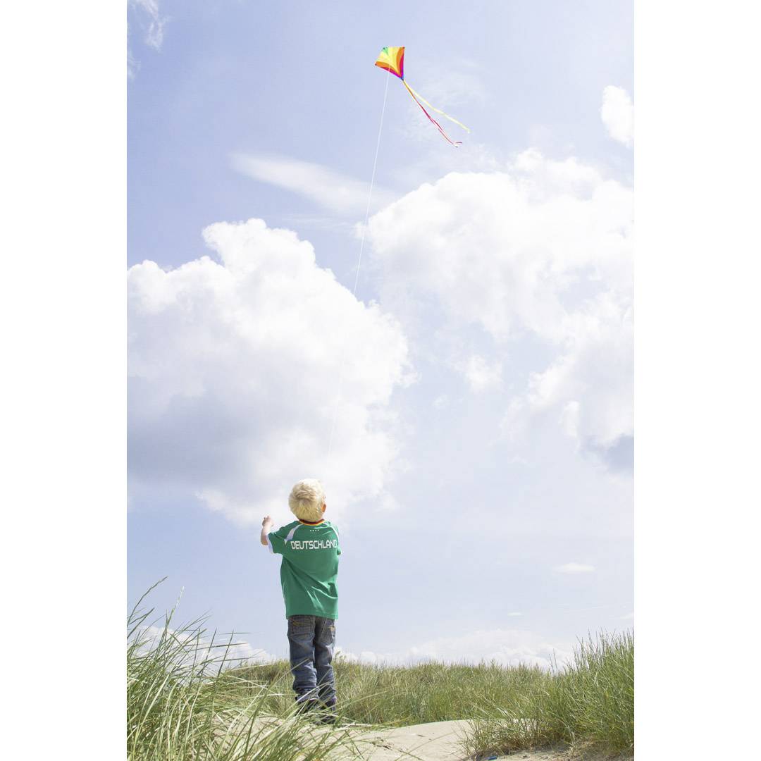 Un enfant portant une veste verte fait voler un cerf-volant multicolore. Un ciel nuageux en arrière-plan, de l'herbe au premier plan.
