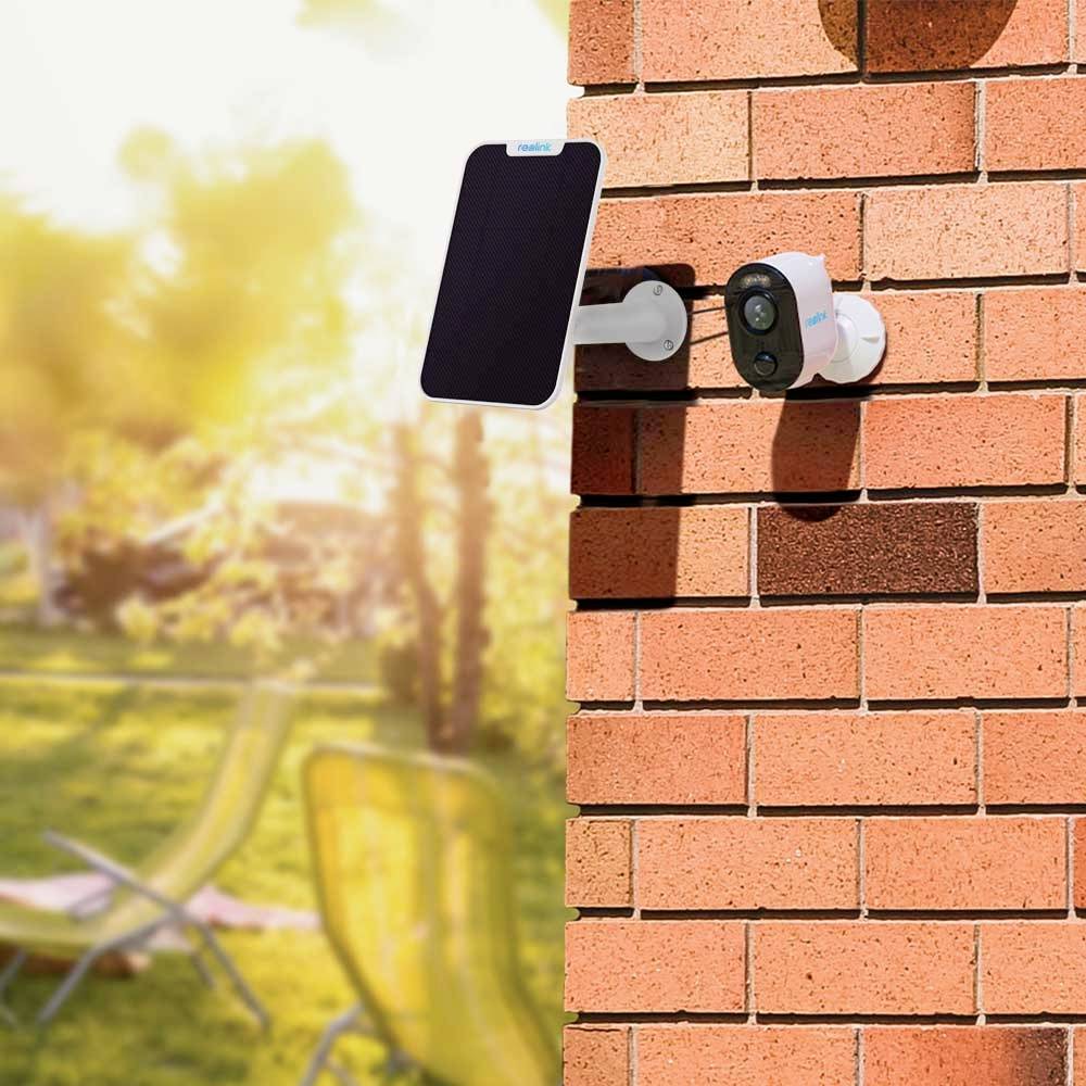 Une caméra de surveillance montée sur un mur de briques et connectée à un panneau solaire. À l'arrière-plan, un jardin flou est visible.