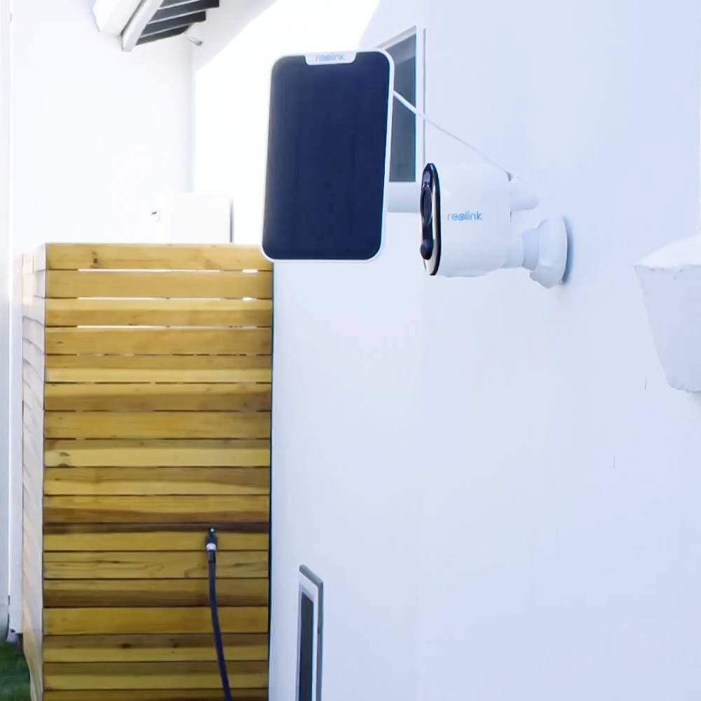 Une caméra de surveillance est installée sur le mur blanc d'un bâtiment. À côté, on peut voir un habillage en bois.