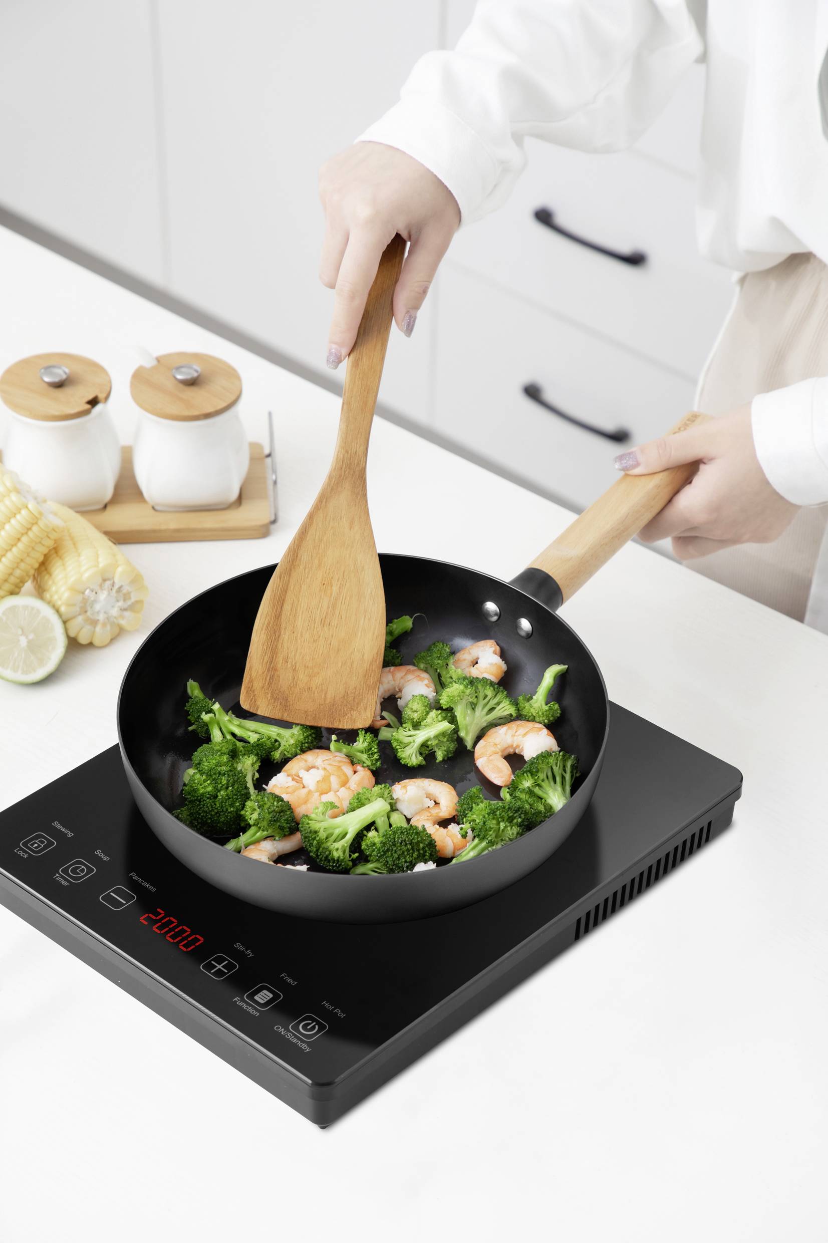 Une personne fait sauter des crevettes et du brocoli dans une poêle sur une plaque à induction. À côté se trouvent des salières et des épis de maïs.
