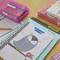Des fournitures scolaires sont posées sur une table : des cahiers, des stylos dans une boîte, ainsi qu'un agenda scolaire avec une chouette sur la couverture.