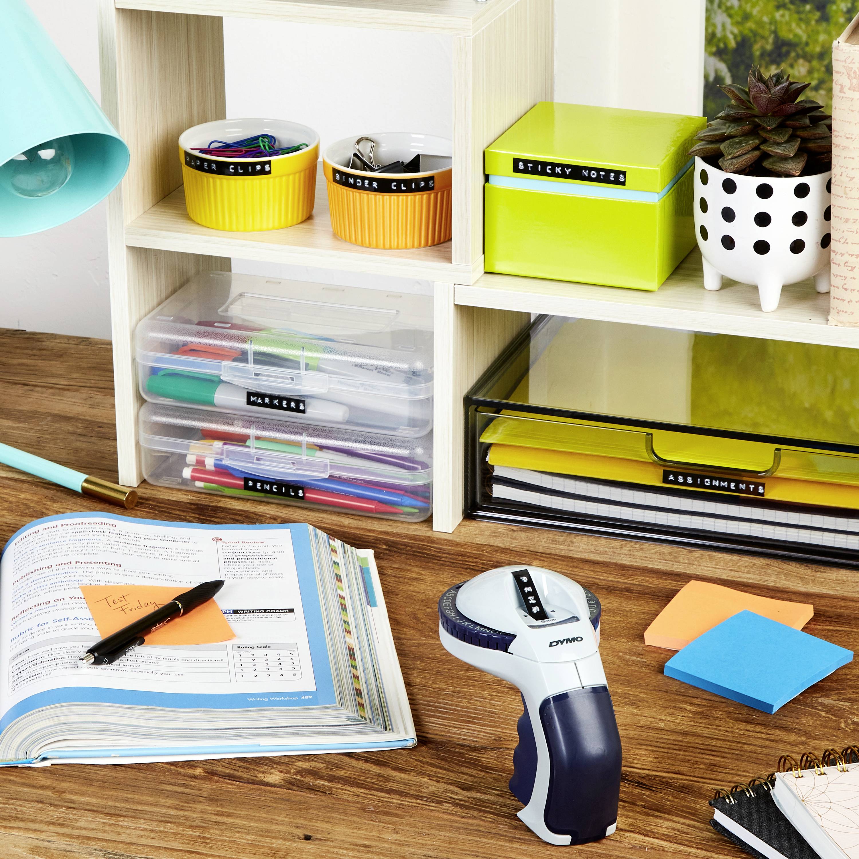 Bureau avec un livre ouvert, un étiqueteur, des stylos, des notes adhésives et un placard contenant du matériel de travail. Décoration discrète.