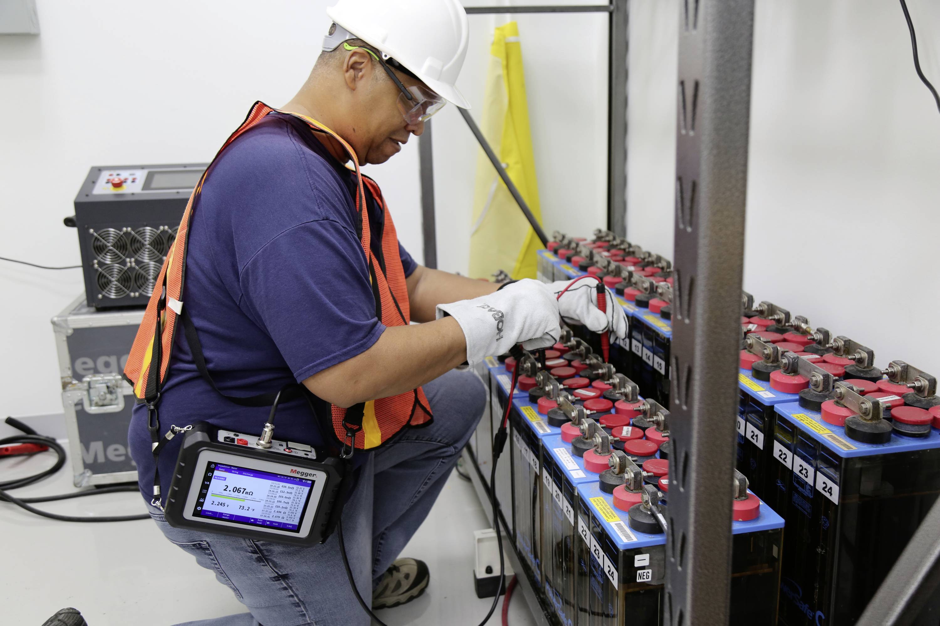 Un technicien portant un équipement de sécurité vérifie des batteries à l'aide d'un appareil de mesure dans un local technique.