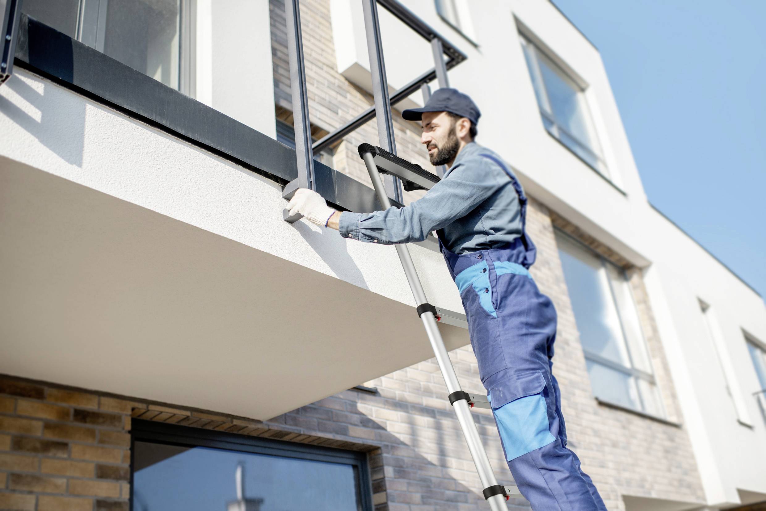Un ouvrier en tenue de travail bleue et casquette est debout sur une échelle et installe une fenêtre de balcon sur un bâtiment moderne.