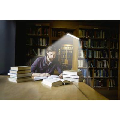 Une personne lit dans une bibliothèque, entourée de livres empilés. Une lampe éclaire la table.
