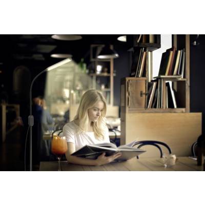 Une femme lit dans un café chaleureux. Un verre est posé sur la table. Des étagères de livres sont visibles en arrière-plan.