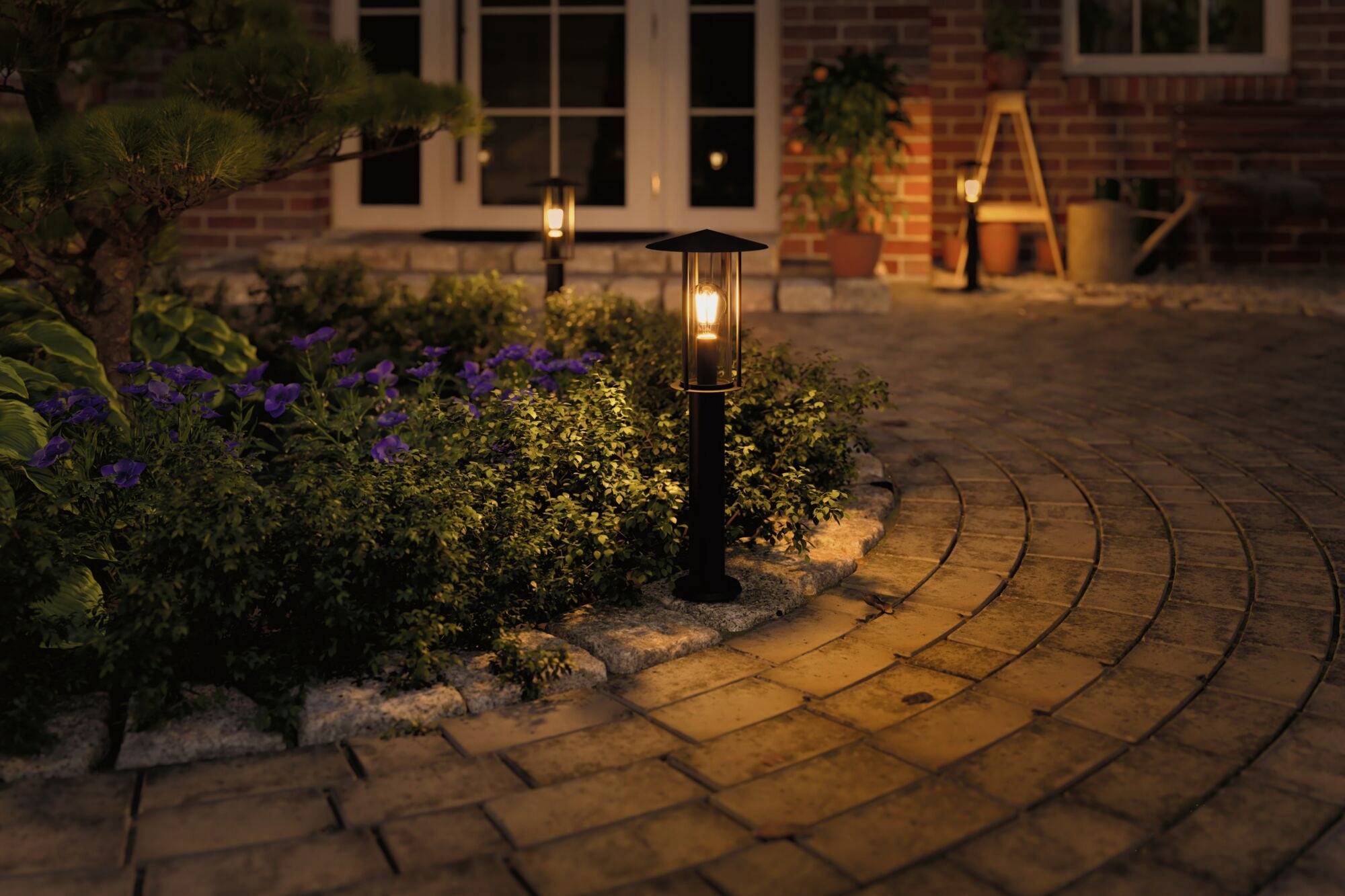 Chemin de jardin pavé la nuit, éclairé par des lanternes, entouré de plantes en fleurs devant une maison aux murs de briques.