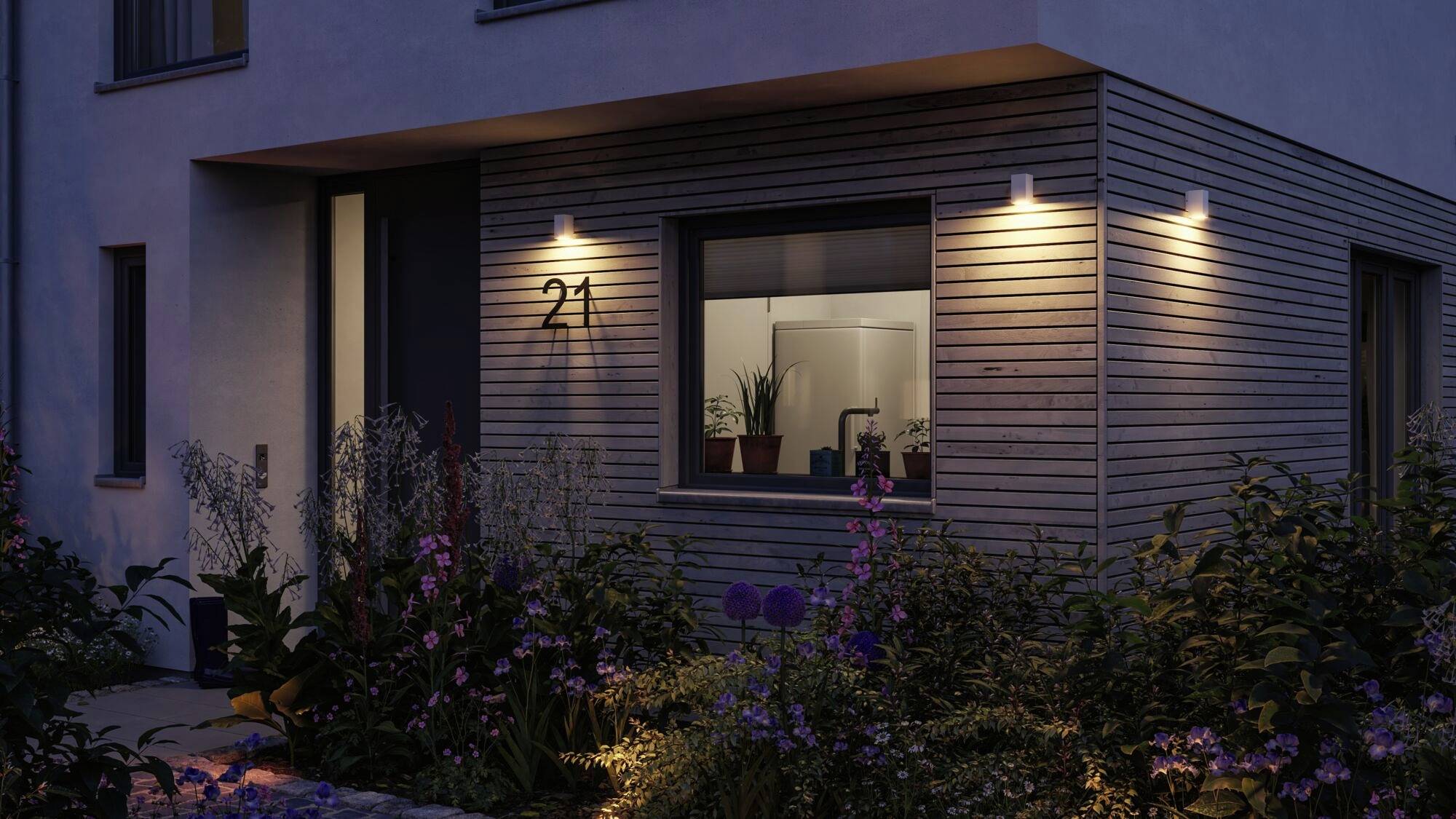 Façade moderne d'une maison au crépuscule avec un hall d'entrée éclairé et le numéro 21. Des plantes bordent le chemin menant à la porte.