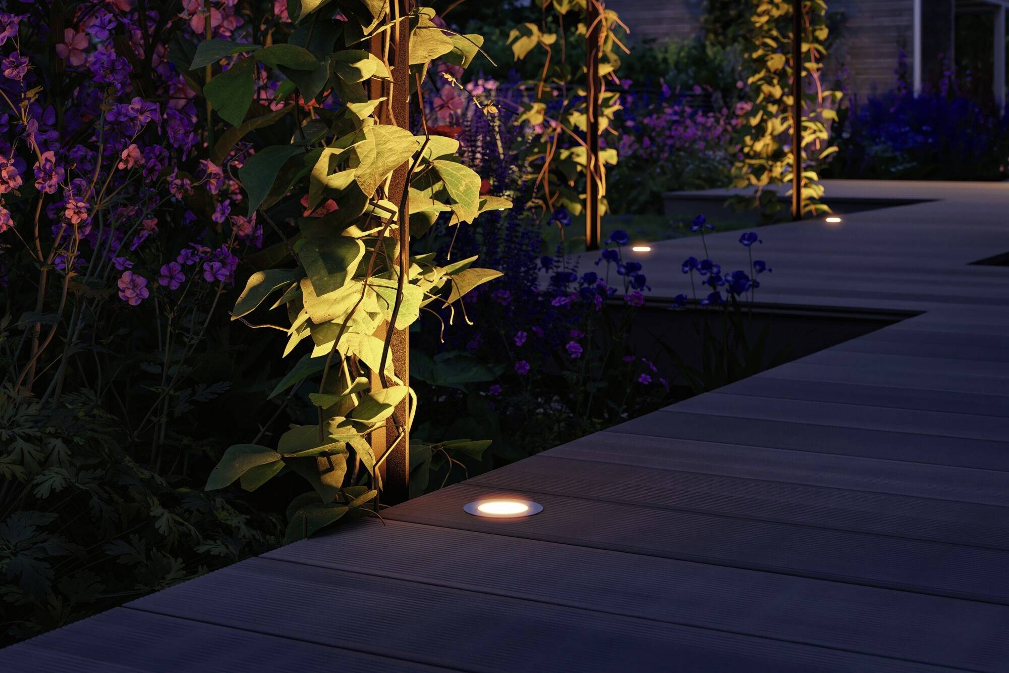 Un chemin de jardin éclairé avec des planches de bois, bordé de plantes vertes et de fleurs illuminées par des lumières chaleureuses.