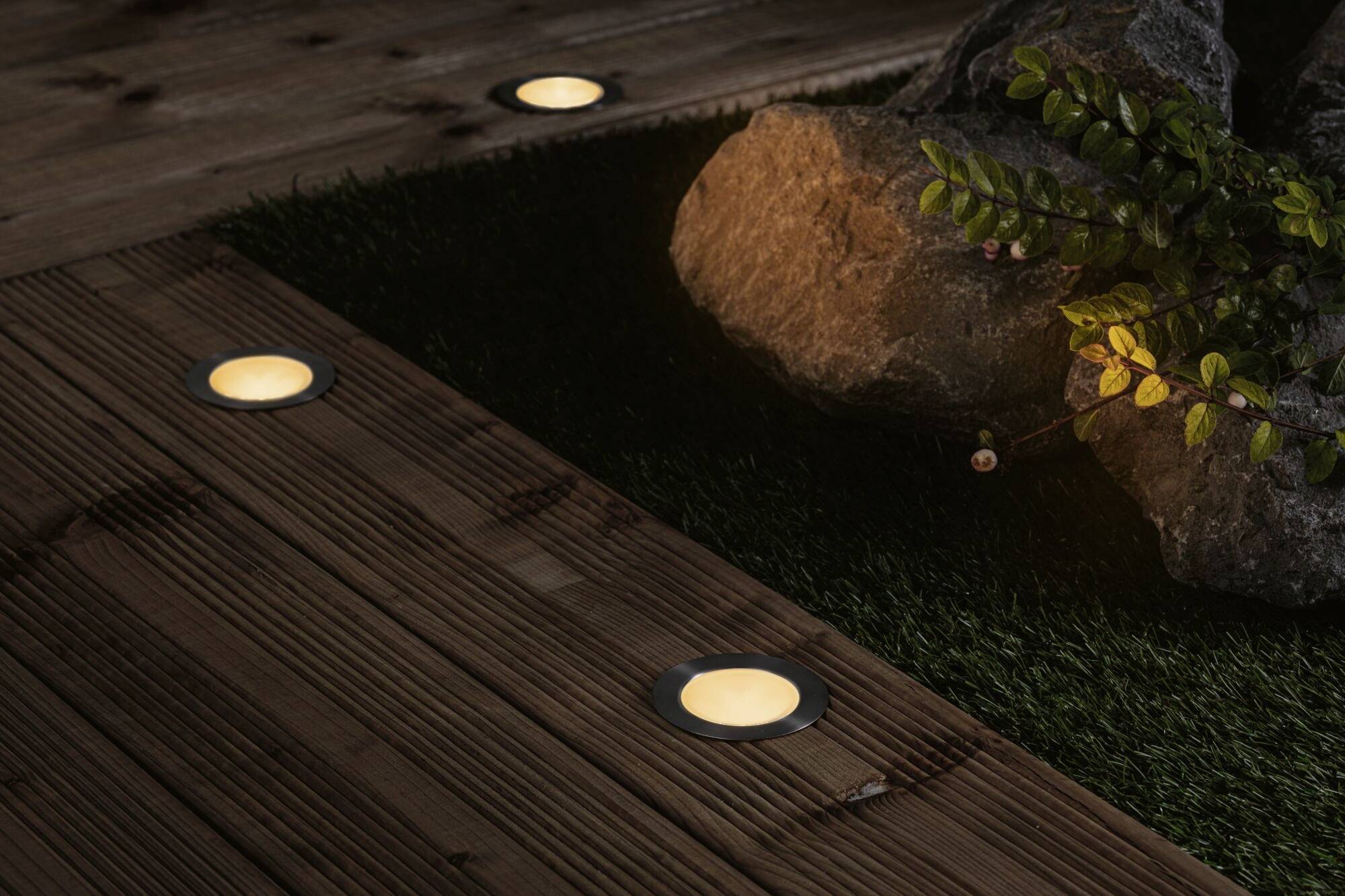 Terrasse en bois avec éclairage intégré, lampes rondes illuminées, pierres décoratives et plante sur gazon artificiel au-dessus.