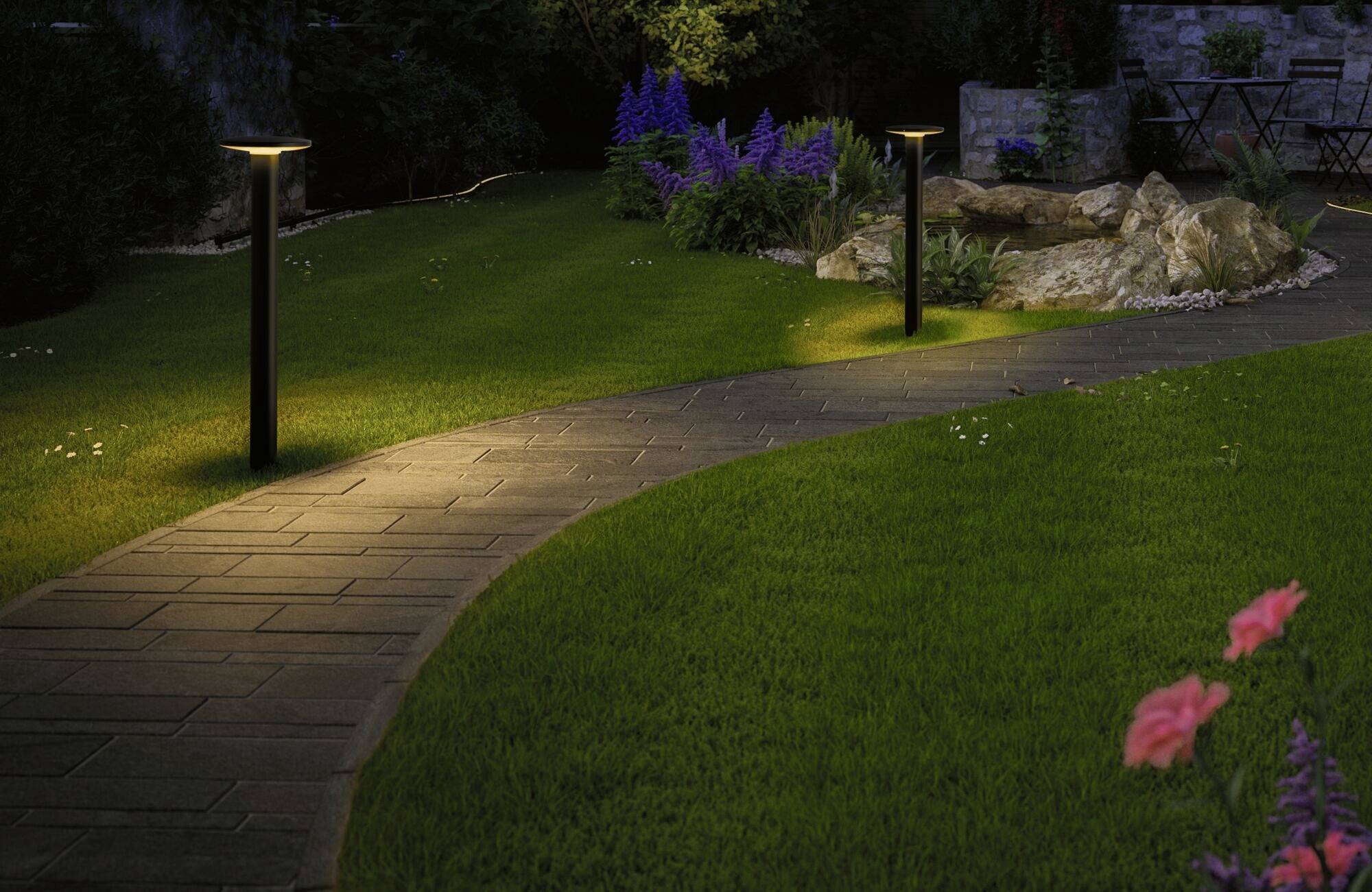 Un chemin de jardin éclairé par des lanternes la nuit, bordé de pelouse soignée et de parterres de fleurs, menant à un jardin de pierres.
