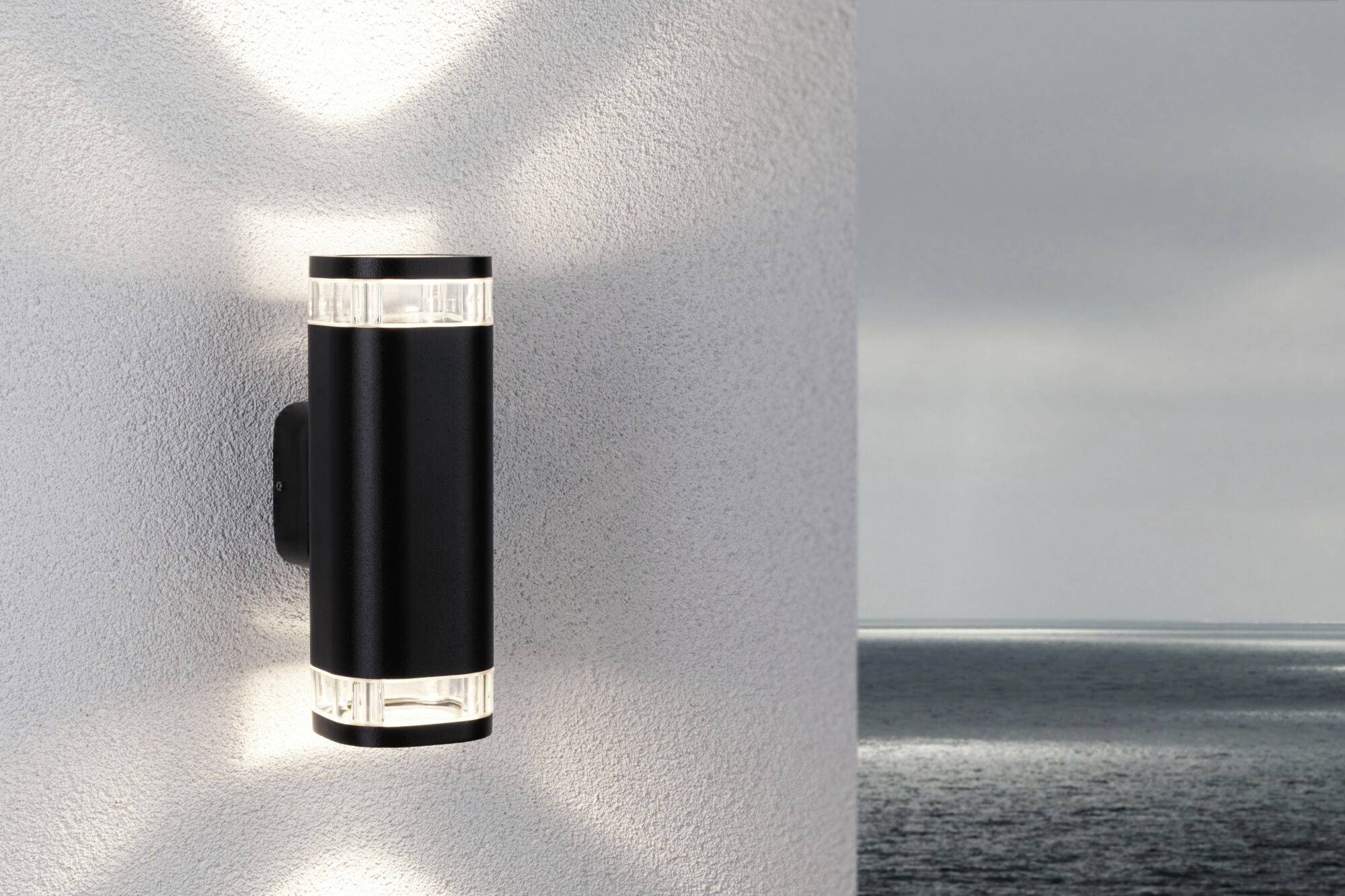 Un applique murale moderne noir émet de la lumière dans les deux sens sur un mur extérieur blanc ; la mer est visible en arrière-plan.