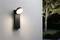 Luminaire extérieur fixé au mur, projetant sa lumière vers le bas, illuminant légèrement les alentours. Un arbre et une pelouse sont visibles en arrière-plan.