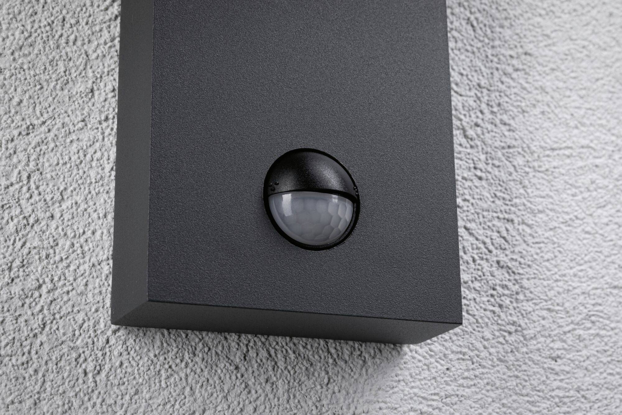 Un détecteur de mouvement noir est installé sur un mur gris à texture structurée.