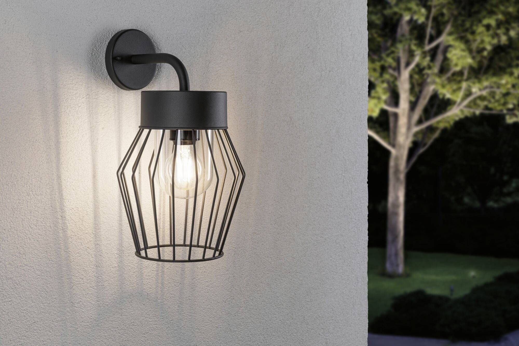 Un luminaire mural moderne en métal noir diffuse une lumière chaleureuse et illumine un paysage arboré à proximité au crépuscule.
