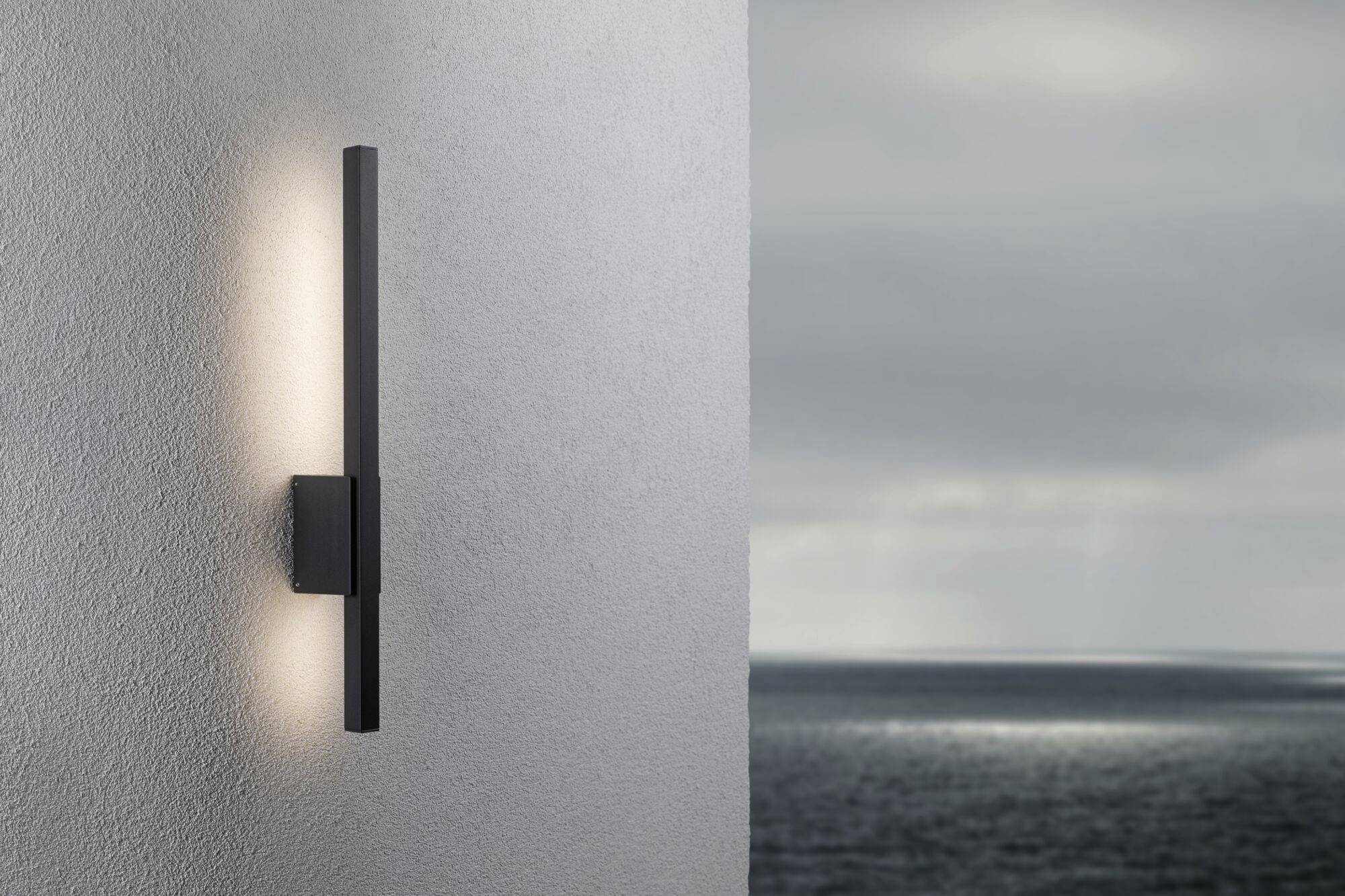Un luminaire mural moderne éclaire un mur en béton. En arrière-plan, un ciel nuageux s'étend au-dessus d'une mer calme.