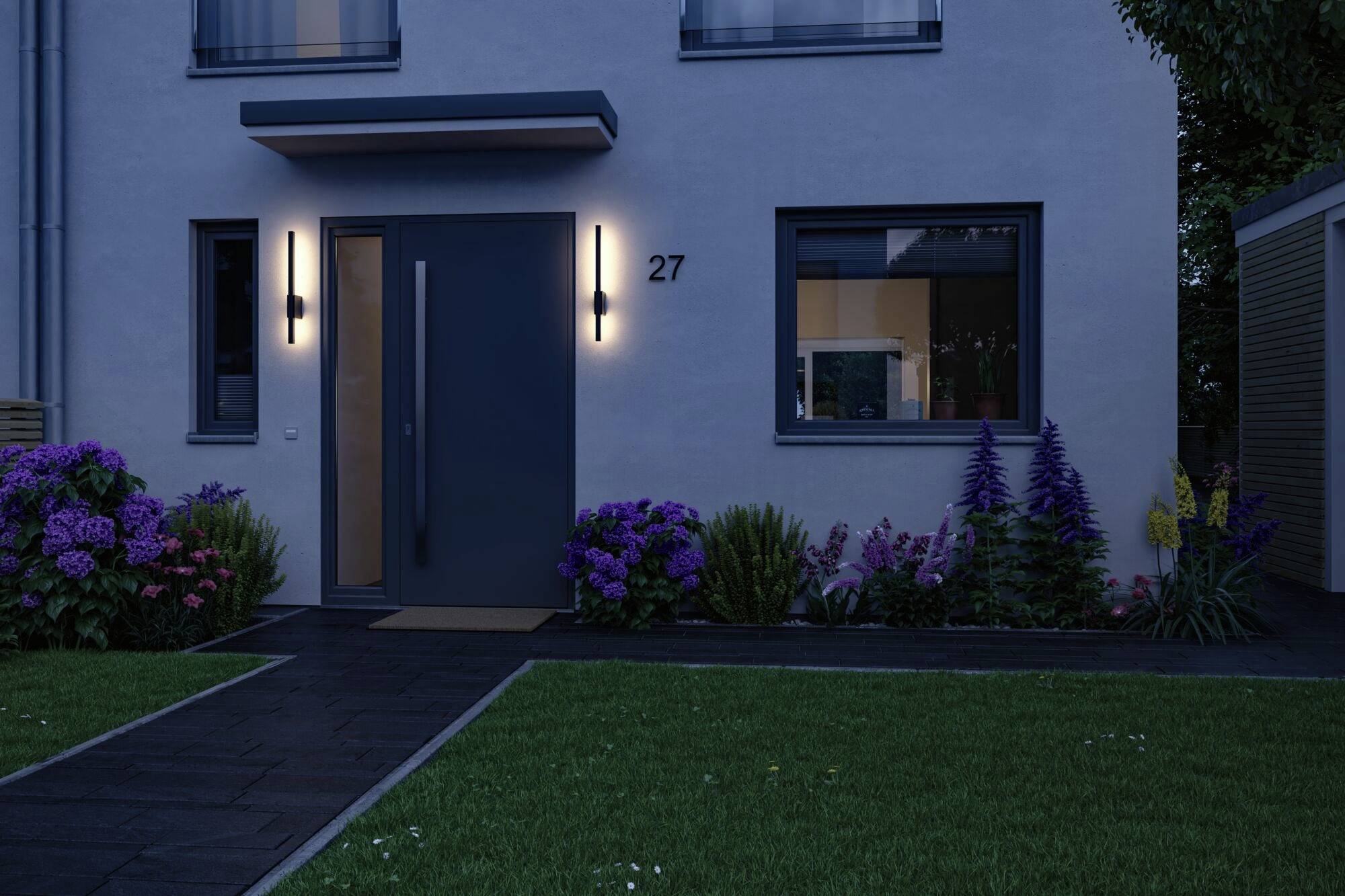 Une maison moderne au crépuscule avec une porte d'entrée illuminée. Entourée de plantes en fleurs devant une pelouse soignée.