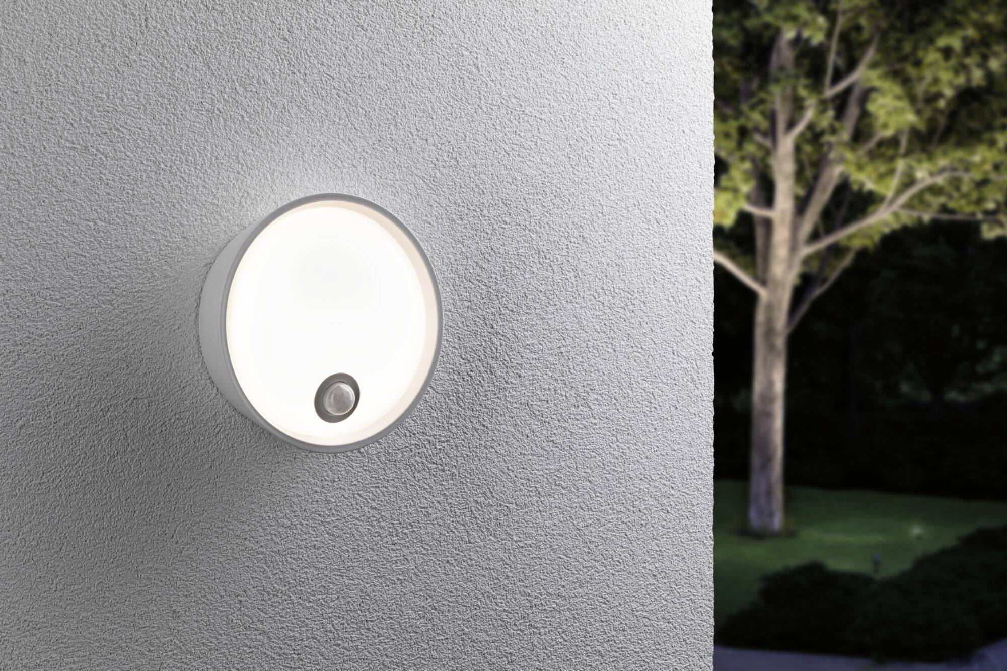 Un luminaire extérieur circulaire avec détecteur de mouvement est fixé sur un mur blanc. En arrière-plan, on aperçoit un arbre au crépuscule.