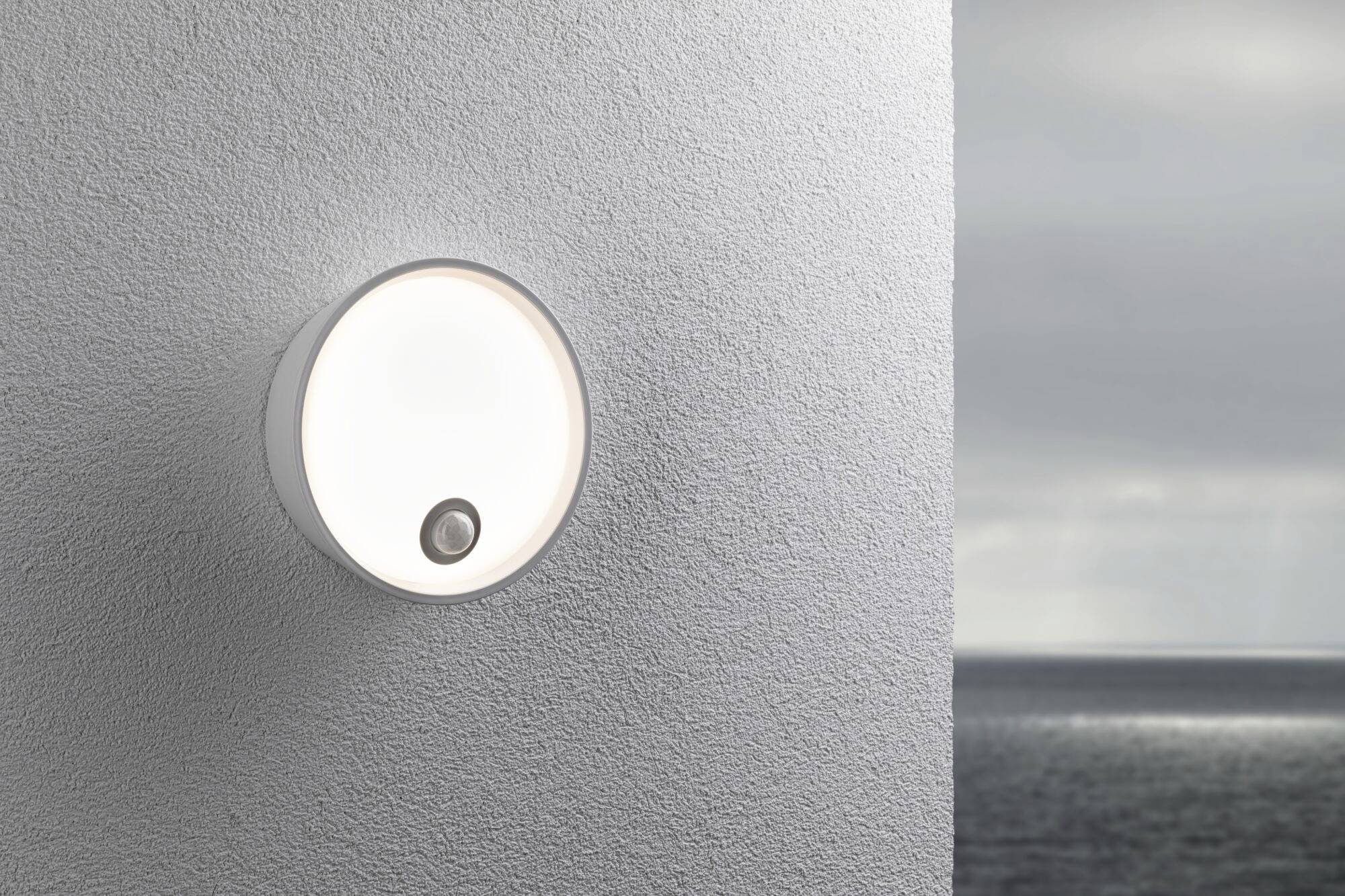 Un luminaire rond et moderne sur un mur blanc avec une vue sur la mer en arrière-plan. La lampe diffuse une lumière douce et uniforme.