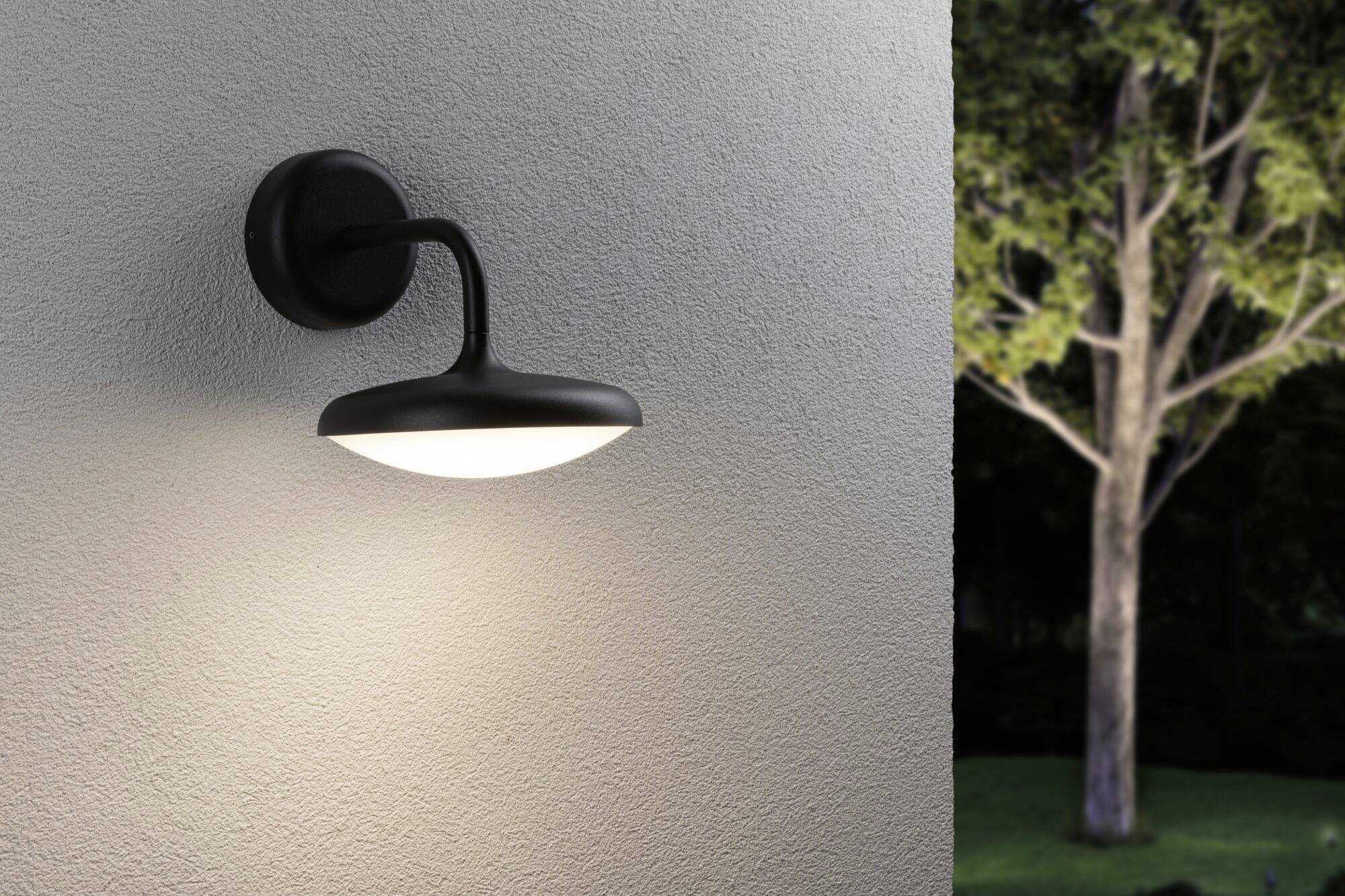 Une lampe murale extérieure éclaire un mur gris. Un arbre est visible en arrière-plan.
