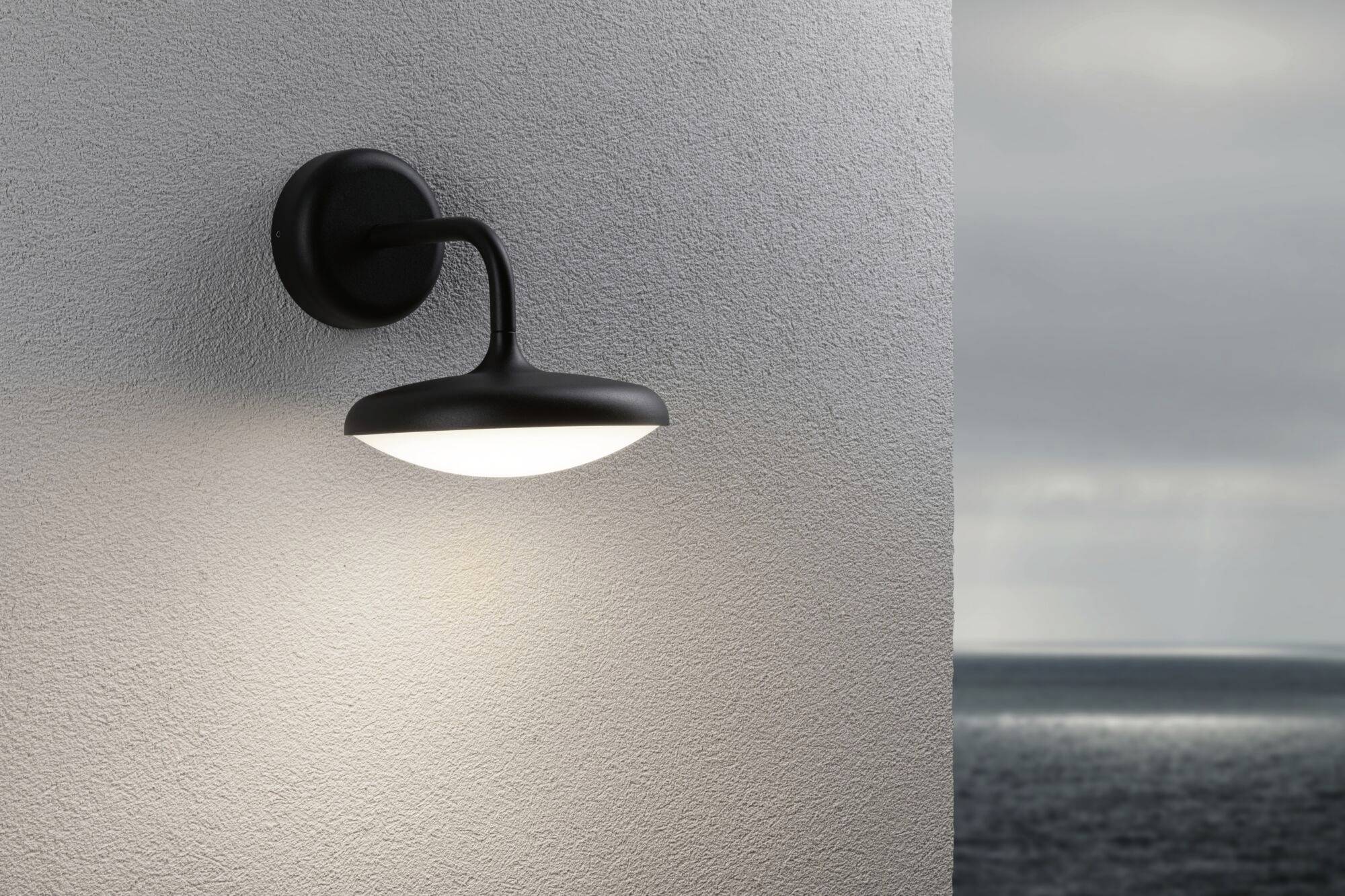 Une lampe murale extérieure brille sur un mur blanc. En arrière-plan, on aperçoit un ciel gris et la mer.
