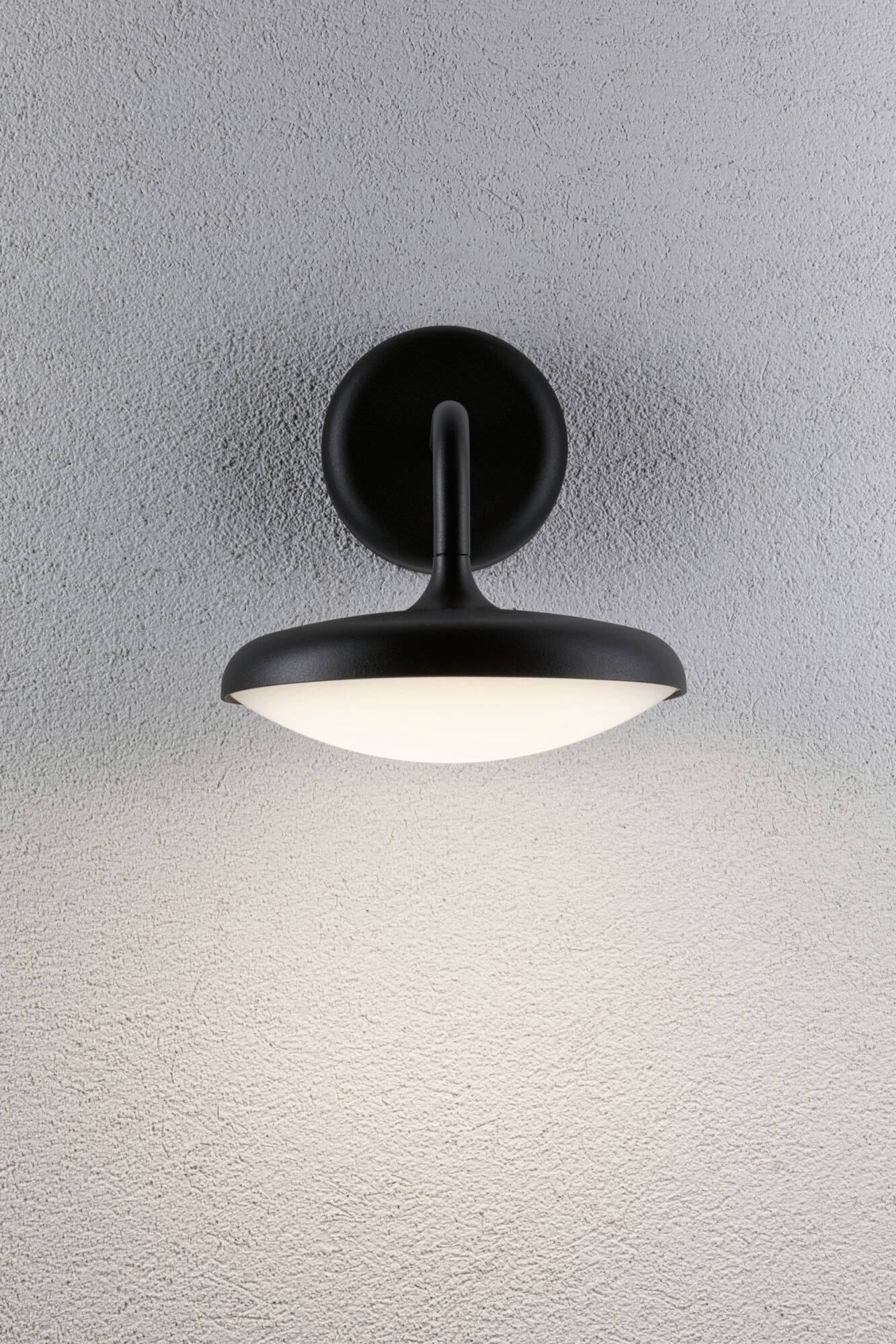Une lampe murale noire diffuse une lumière douce et chaleureuse sur un mur gris, créant une atmosphère accueillante.
