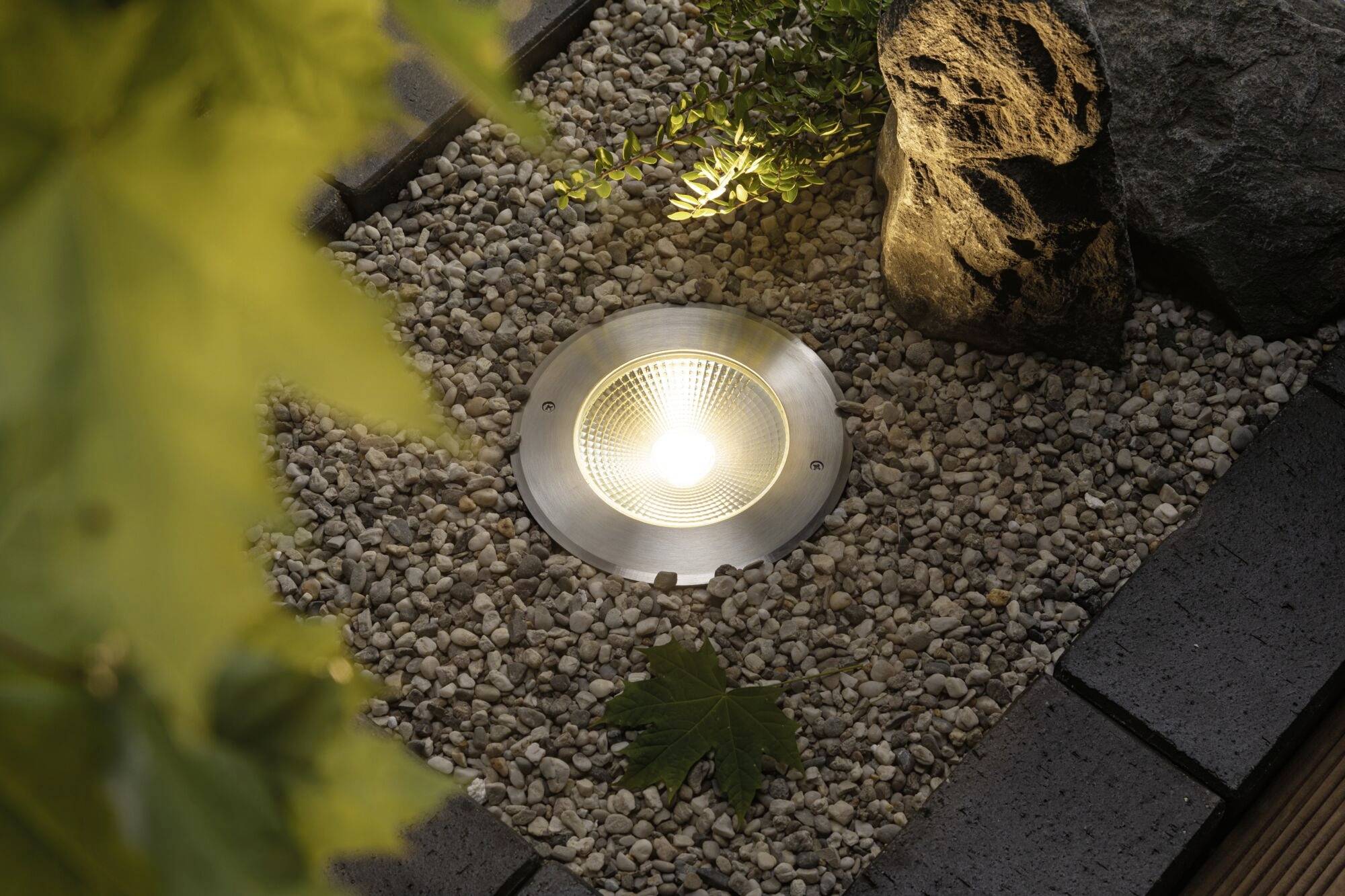 Une lampe est encastrée dans un sol de jardin recouvert de galets, entourée de plantes et d'une pierre, et diffuse une lumière vive.