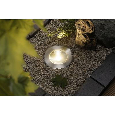 Une lampe est encastrée dans un sol de jardin recouvert de galets, entourée de plantes et d'une pierre, et diffuse une lumière vive.
