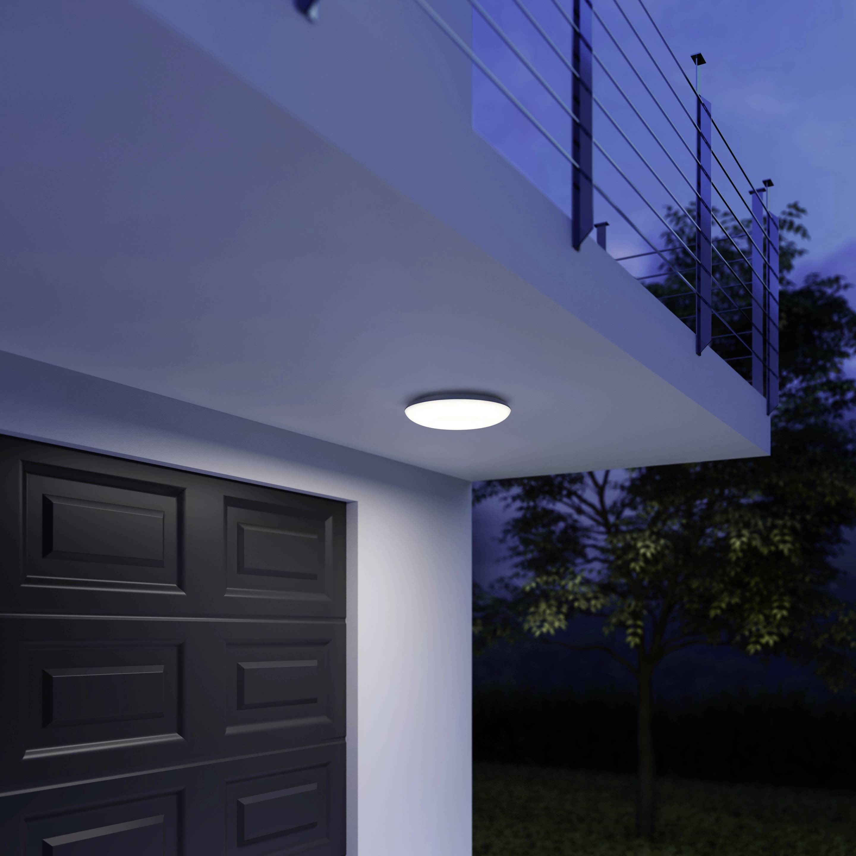 Une maison moderne avec une terrasse d'entrée illuminée et une porte de garage sombre. Des arbres sont visibles en arrière-plan pendant la nuit.