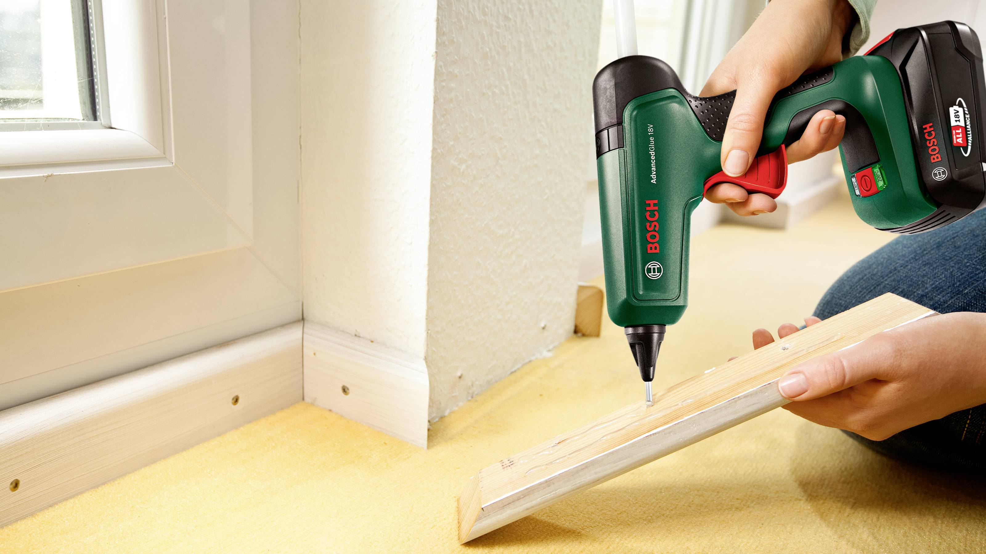 Une personne utilise une visseuse sans fil Bosch pour fixer une latte en bois dans le coin d'une pièce, à côté d'un encadrement de fenêtre.