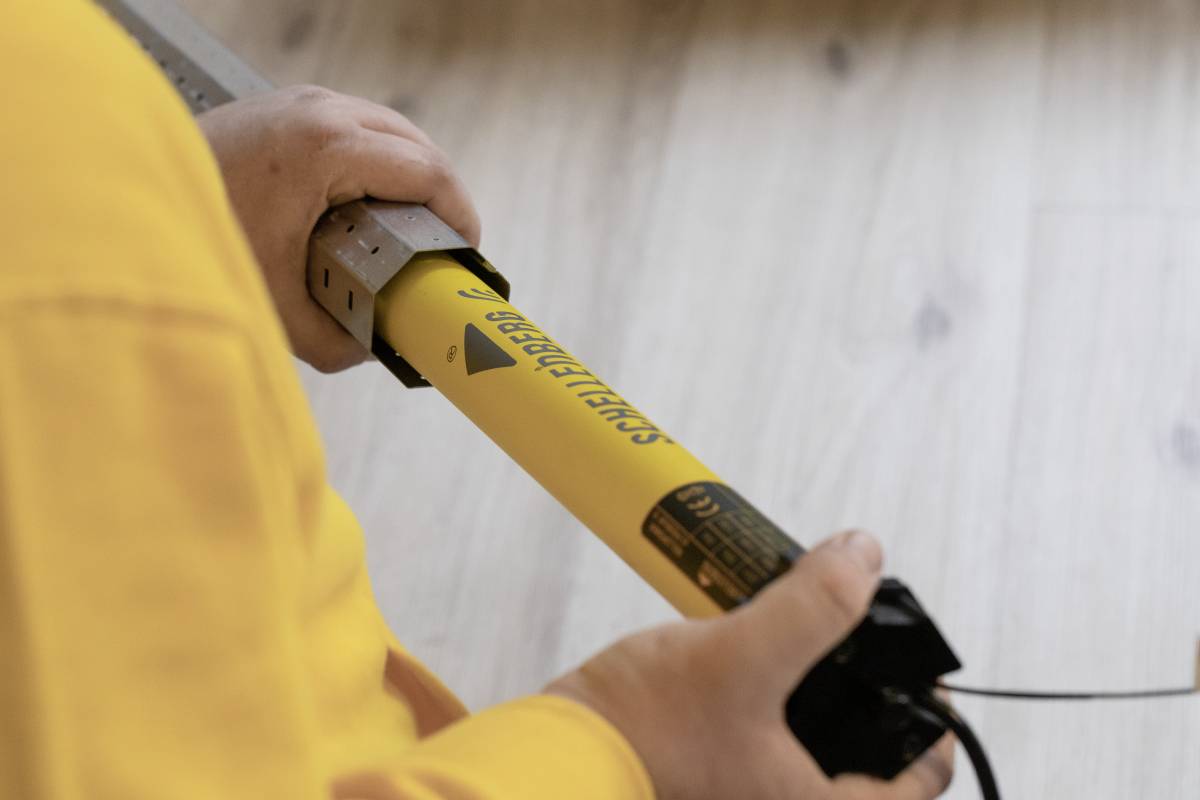 Une personne portant un t-shirt jaune tient un capteur de sécurité à faisceau lumineux étiqueté « SICK », probablement utilisé à des fins industrielles ou de sécurité.