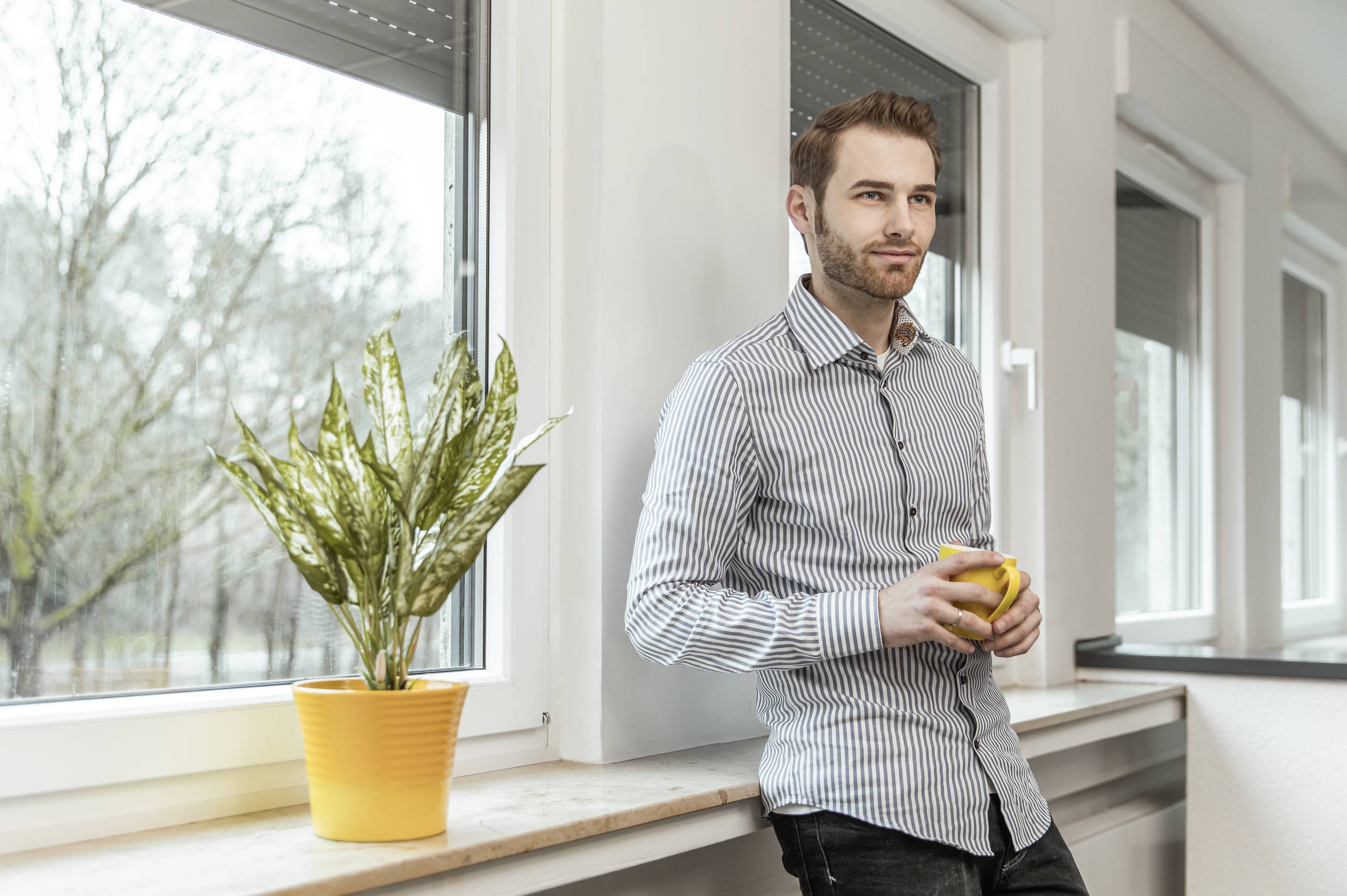 Un homme portant une chemise rayée est adossé à un rebord de fenêtre et tient une tasse jaune. Une plante en pot se trouve à côté de lui.