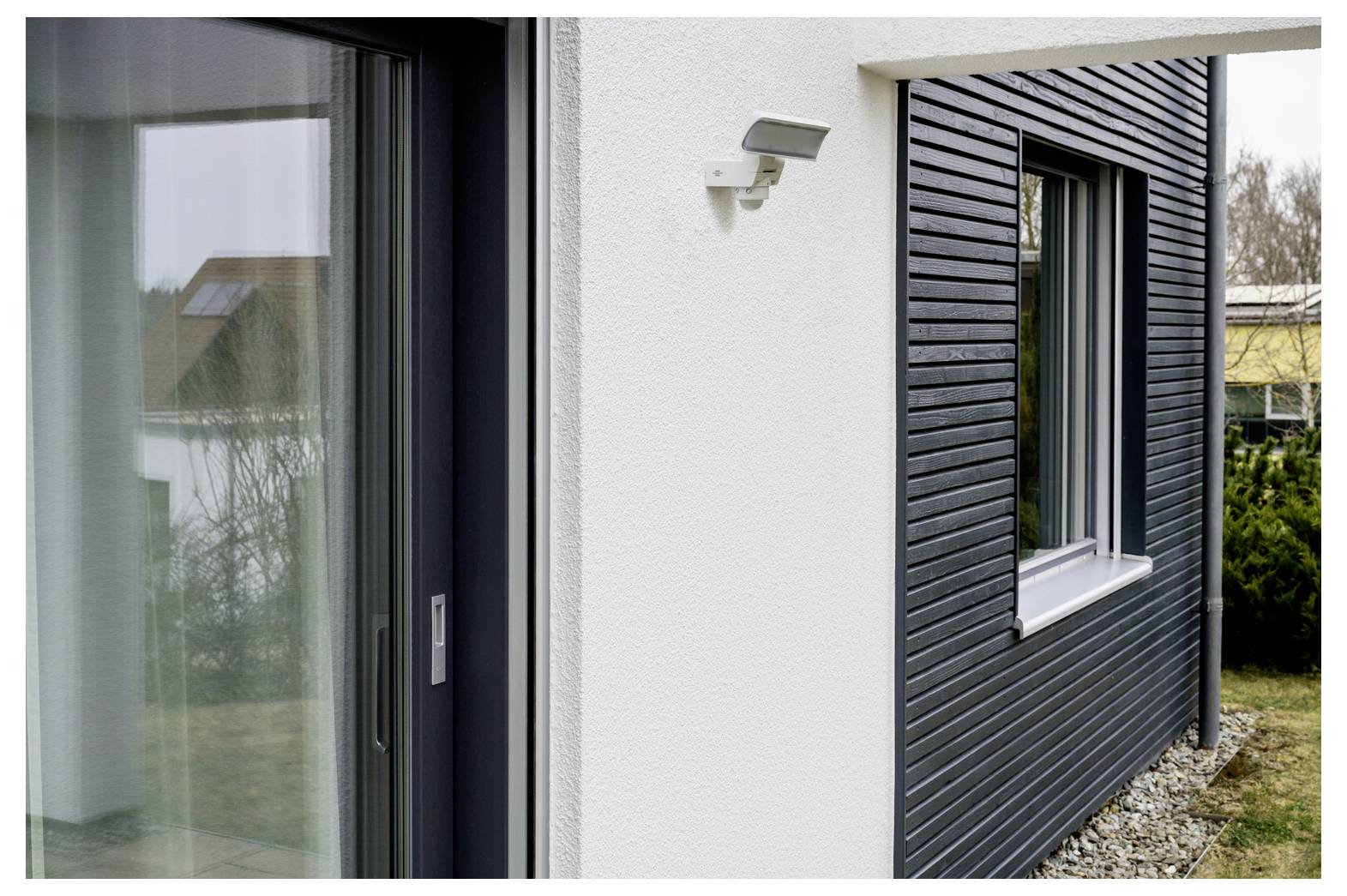Extérieur d'une maison moderne montrant une porte coulissante en verre, un mur en stuc blanc avec applique murale, et un bardage horizontal foncé à côté d'une fenêtre.