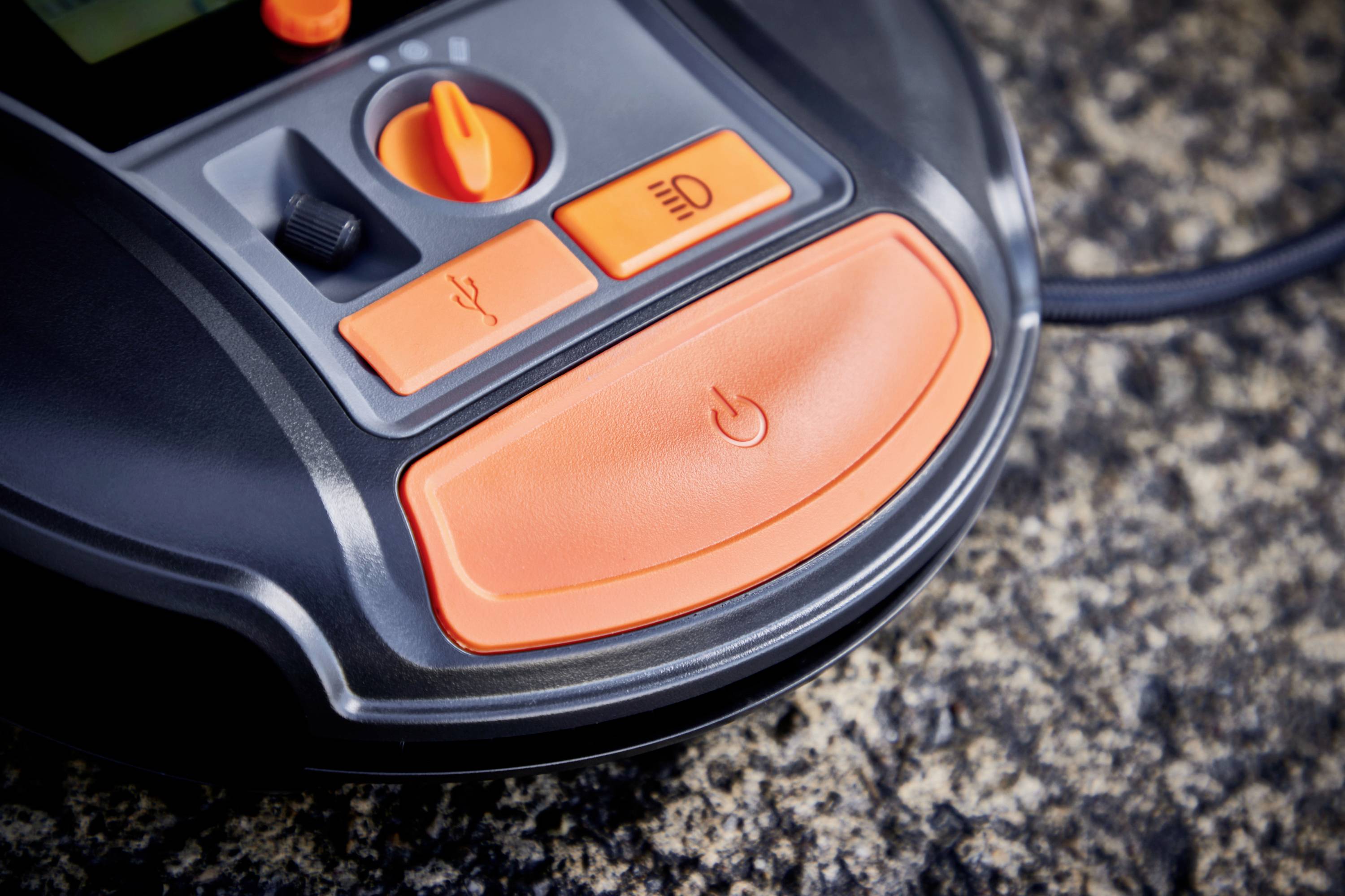 L'image montre un gros plan du panneau de commande d'un Automower avec des touches orange pour différentes fonctions sur une surface en pierre.