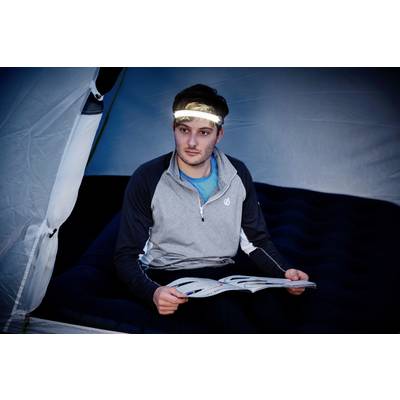 Une personne est assise dans une tente sur un matelas pneumatique et lit un magazine. Elle porte un bandeau frontal avec une lumière qui éclaire son visage.