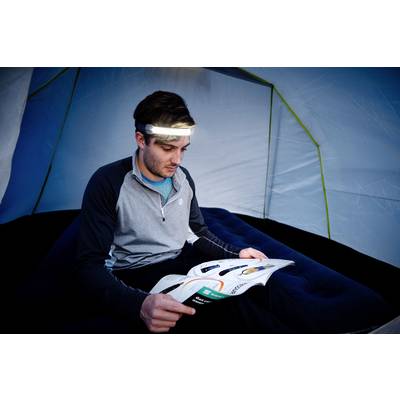 Une personne est assise dans une tente sur un matelas pneumatique, en train de lire un livre avec une lampe frontale sur la tête.