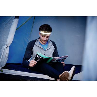 Un homme portant une lampe frontale lit un magazine à l'intérieur d'une tente.