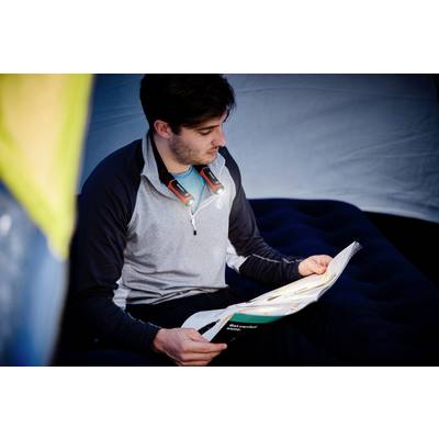 Une personne est assise dans une tente et lit un magazine. Elle porte une lampe autour du cou qui facilite sa lecture.