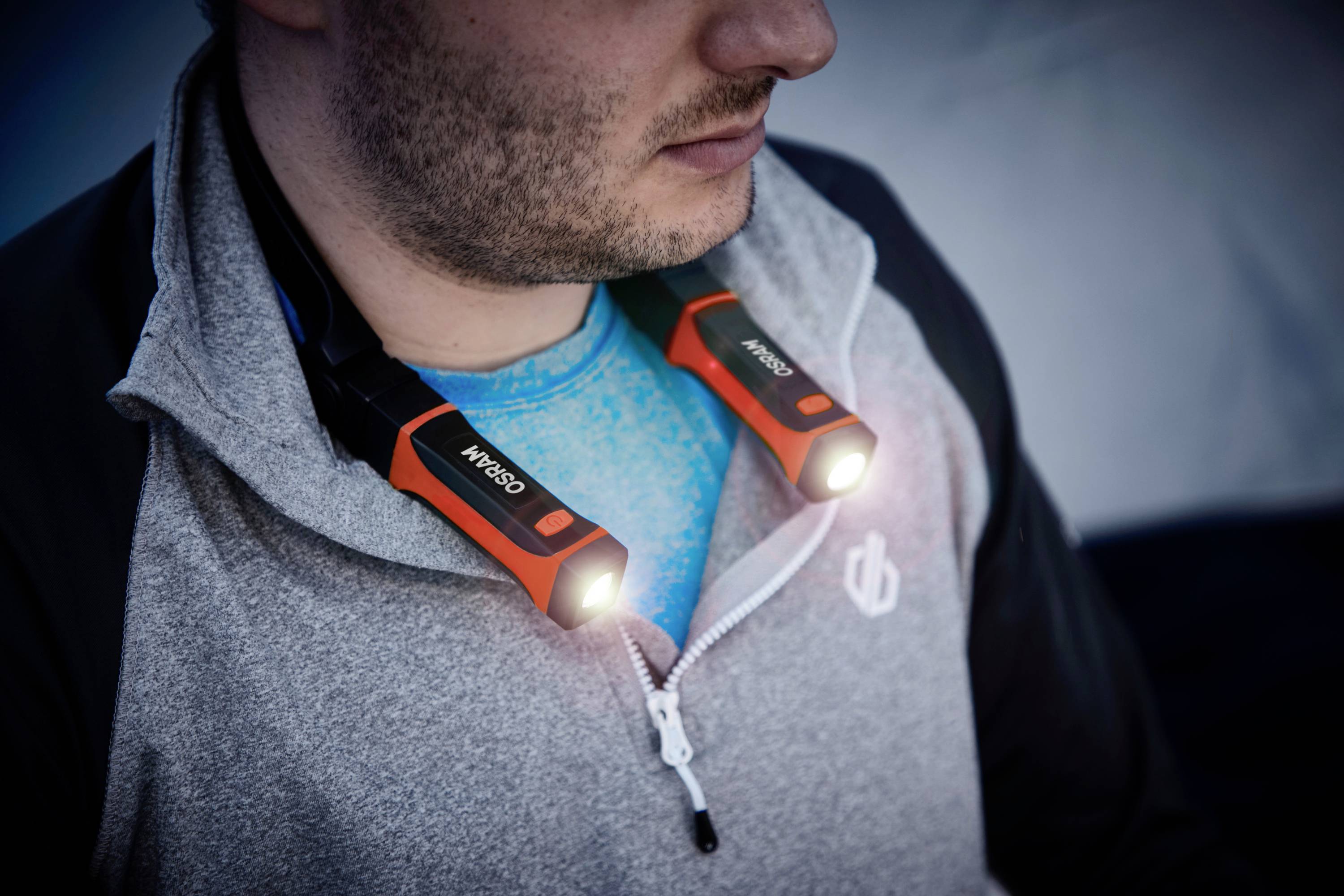 Un homme porte une veste grise et une lampe LED autour du cou. La lampe est orange et brille dans deux directions.