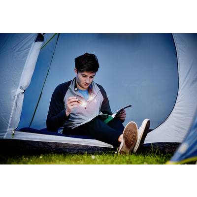 Un homme est assis dans une tente et lit un livre à la lumière d'une lampe de poche. C'est le soir, et il semble concentré.