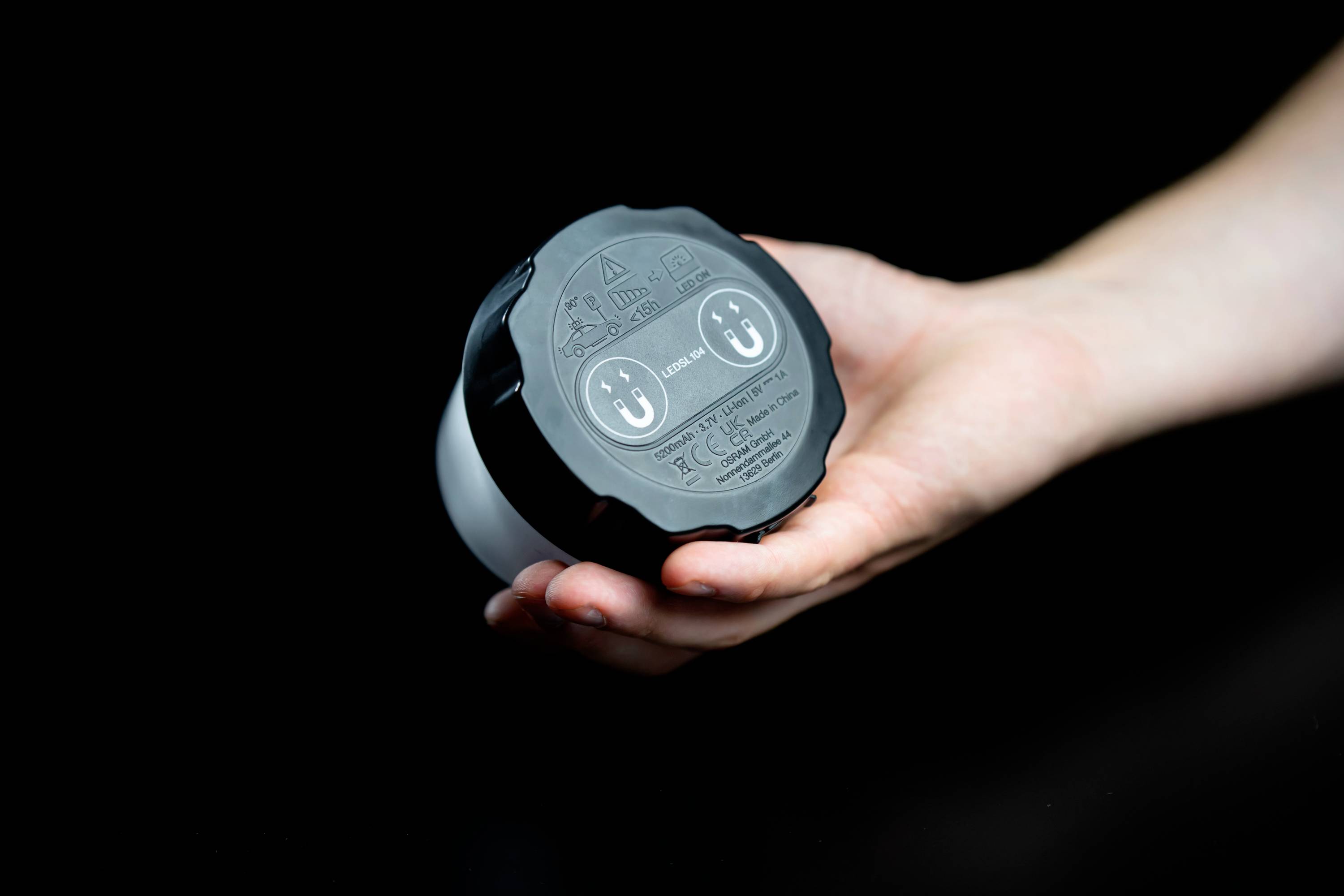 Une main tient un objet rond noir avec deux prises et des symboles sur le dessus. L'arrière-plan est noir.