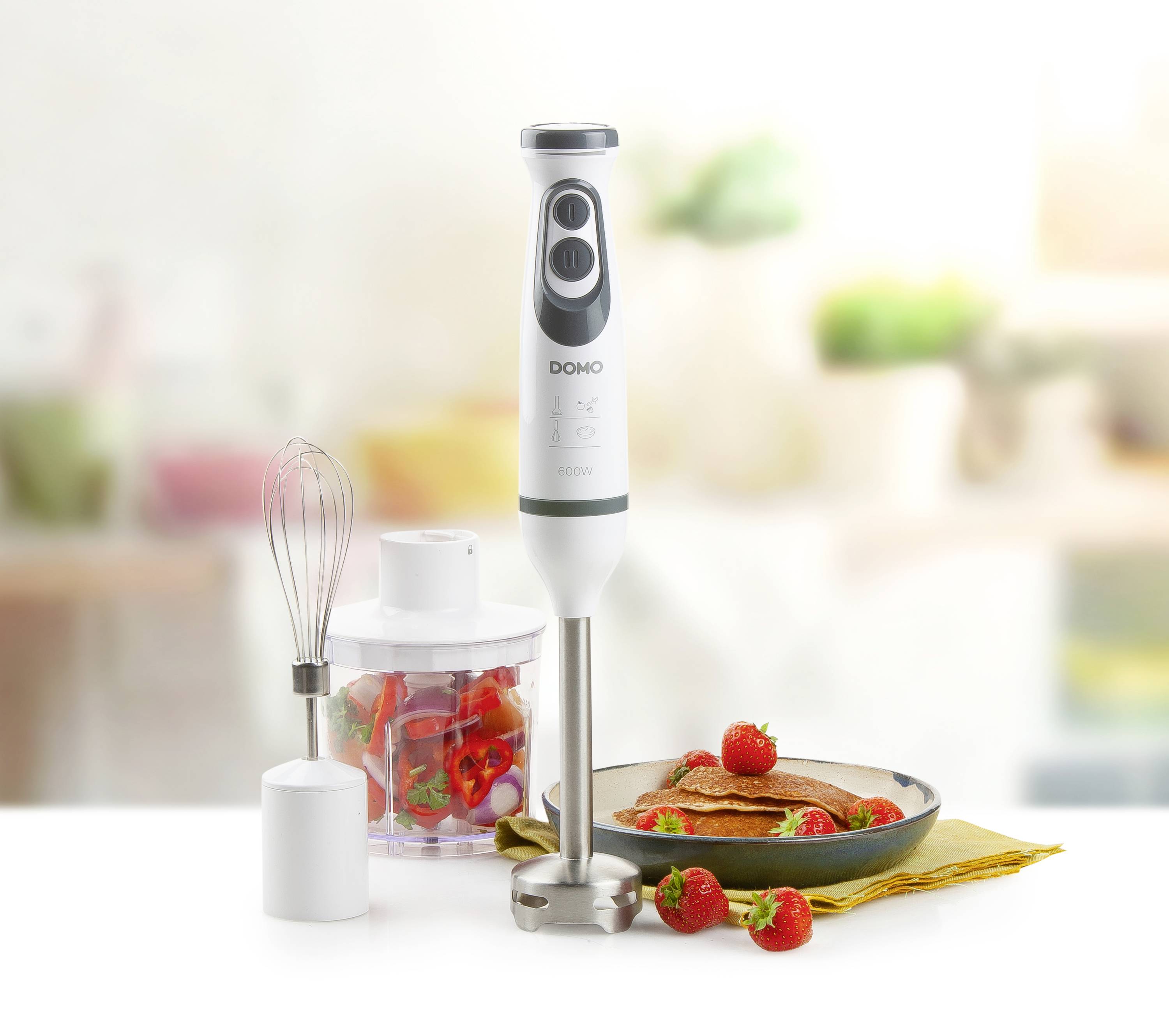 Un mixeur plongeant avec ses accessoires est posé sur une table. À côté, on trouve des fraises fraîches, un fouet et un récipient rempli.