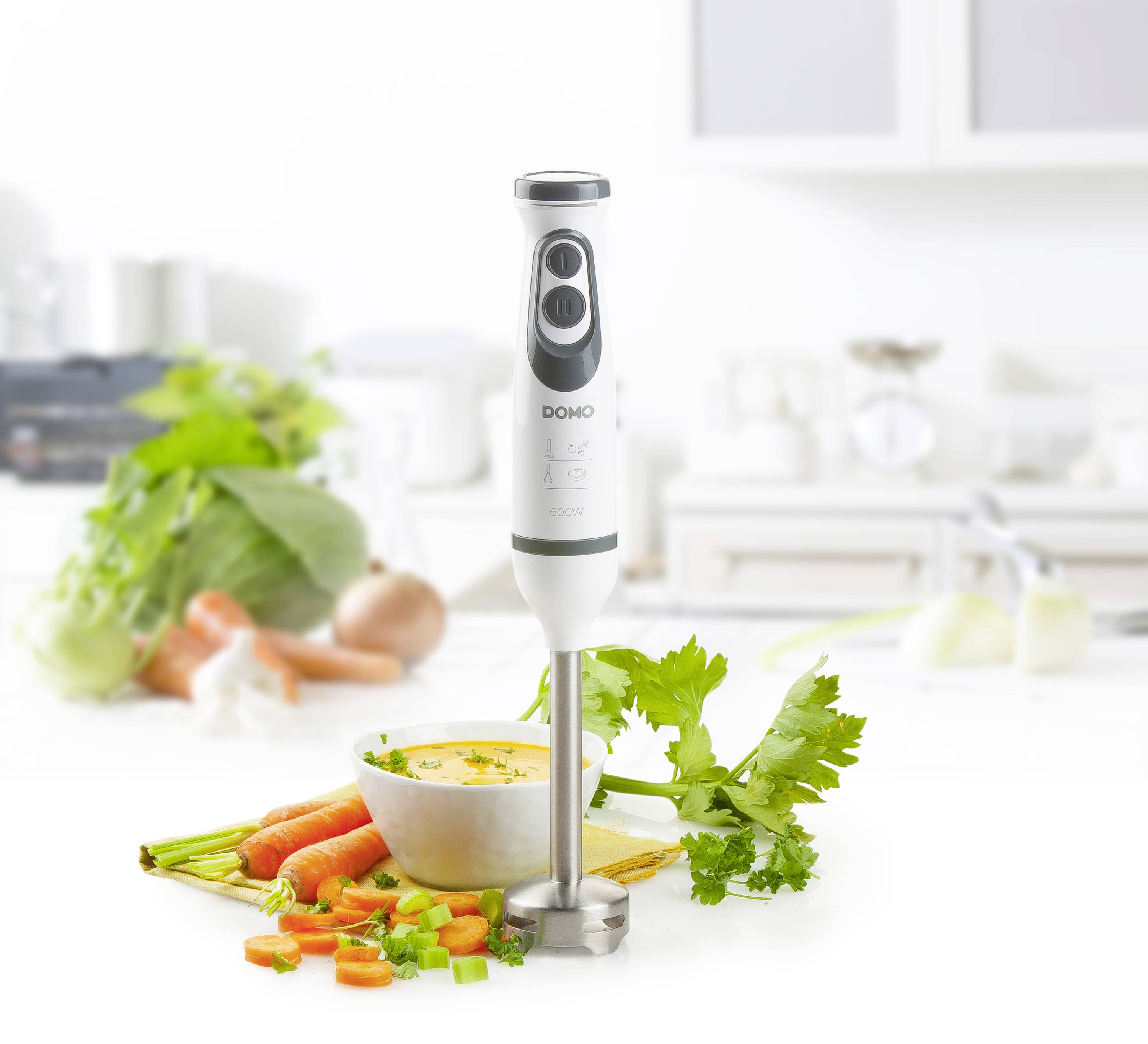 Un mixeur plongeant est posé à côté d'un bol de soupe sur une table de cuisine. Tout autour sont disposés des carottes fraîches, des oignons et du céleri.